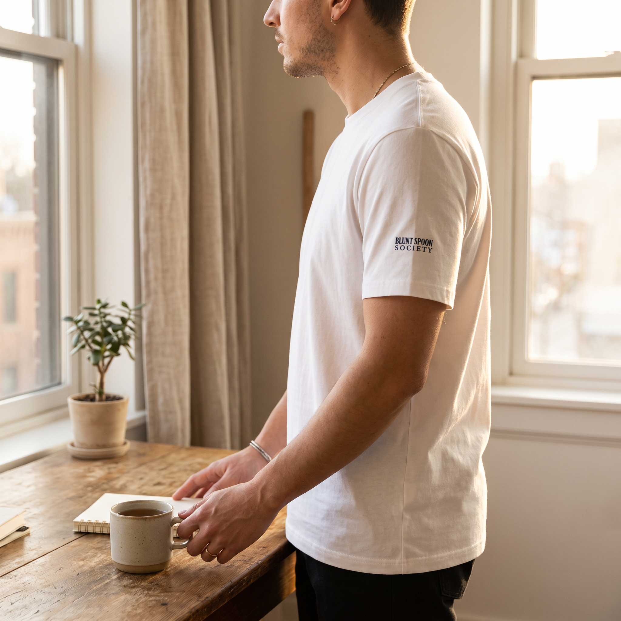 Man in a white Blunt Spoon Society t-shirt standing by a window next to a table with a coffee cup