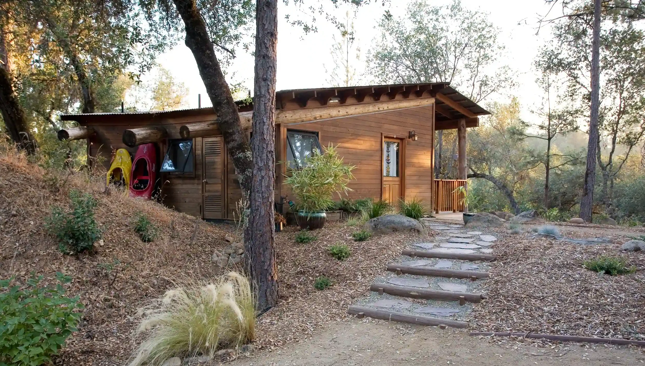 Small wooden cabin on a sloped, wooded landscape with stone pathway, potted plants, and colorful kayaks hanging on the side.