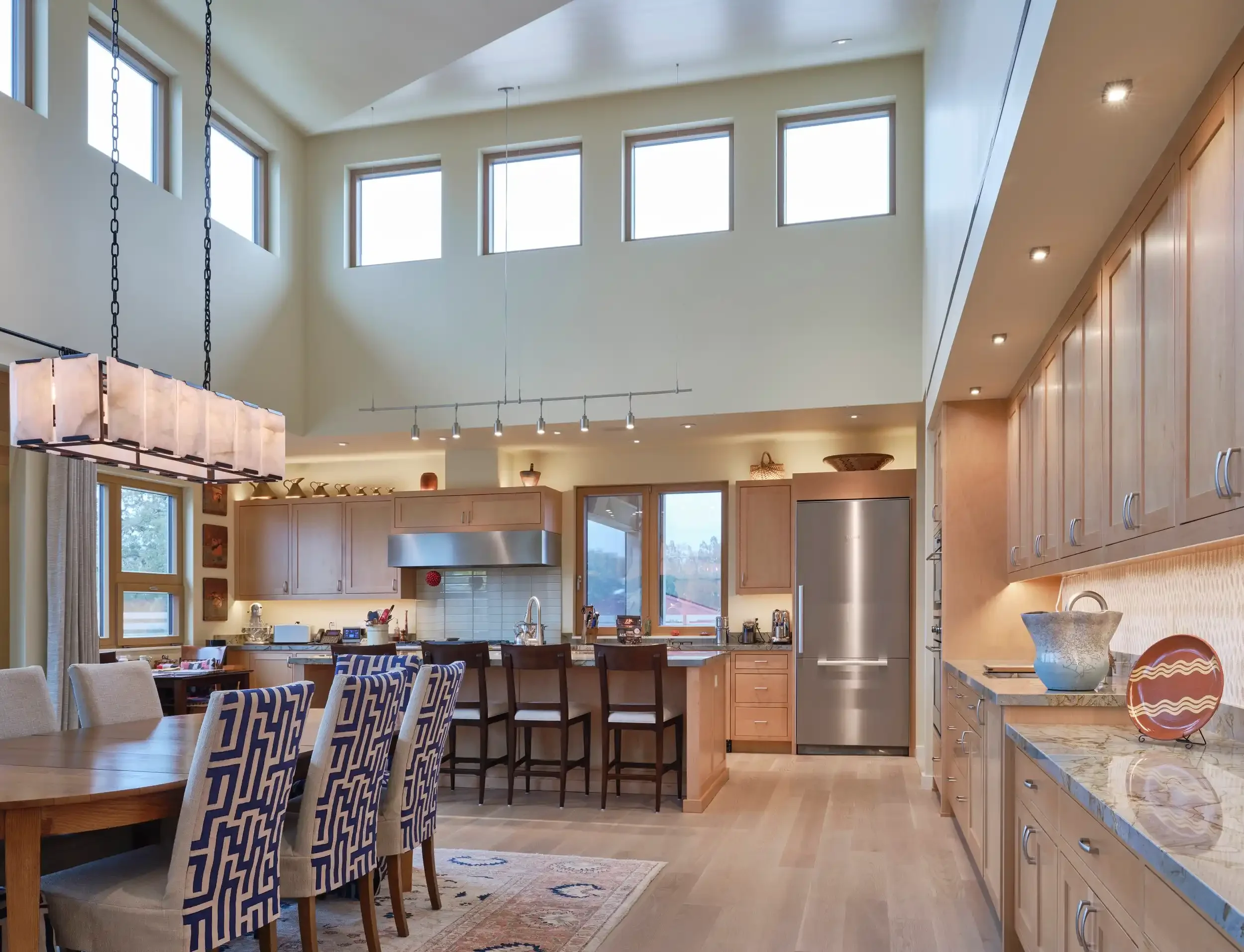 Open-concept kitchen and dining area with high ceilings, large windows, wooden cabinets, stainless steel appliances, and a wooden dining table with patterned chairs.