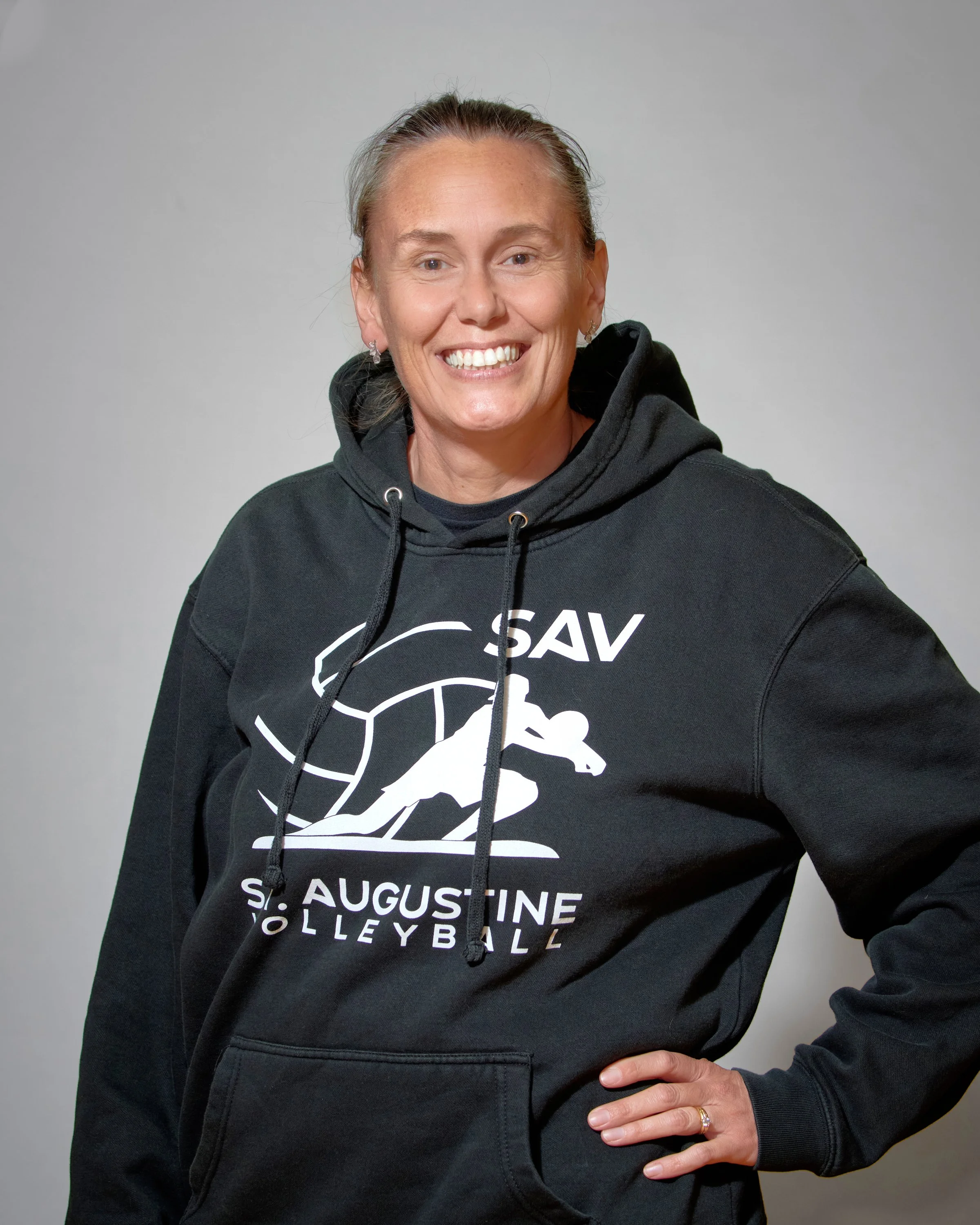 A woman with long red hair smiling at the camera, wearing a dark gray T-shirt with a volleyball graphic and the words "St. Augustine Volleyball."