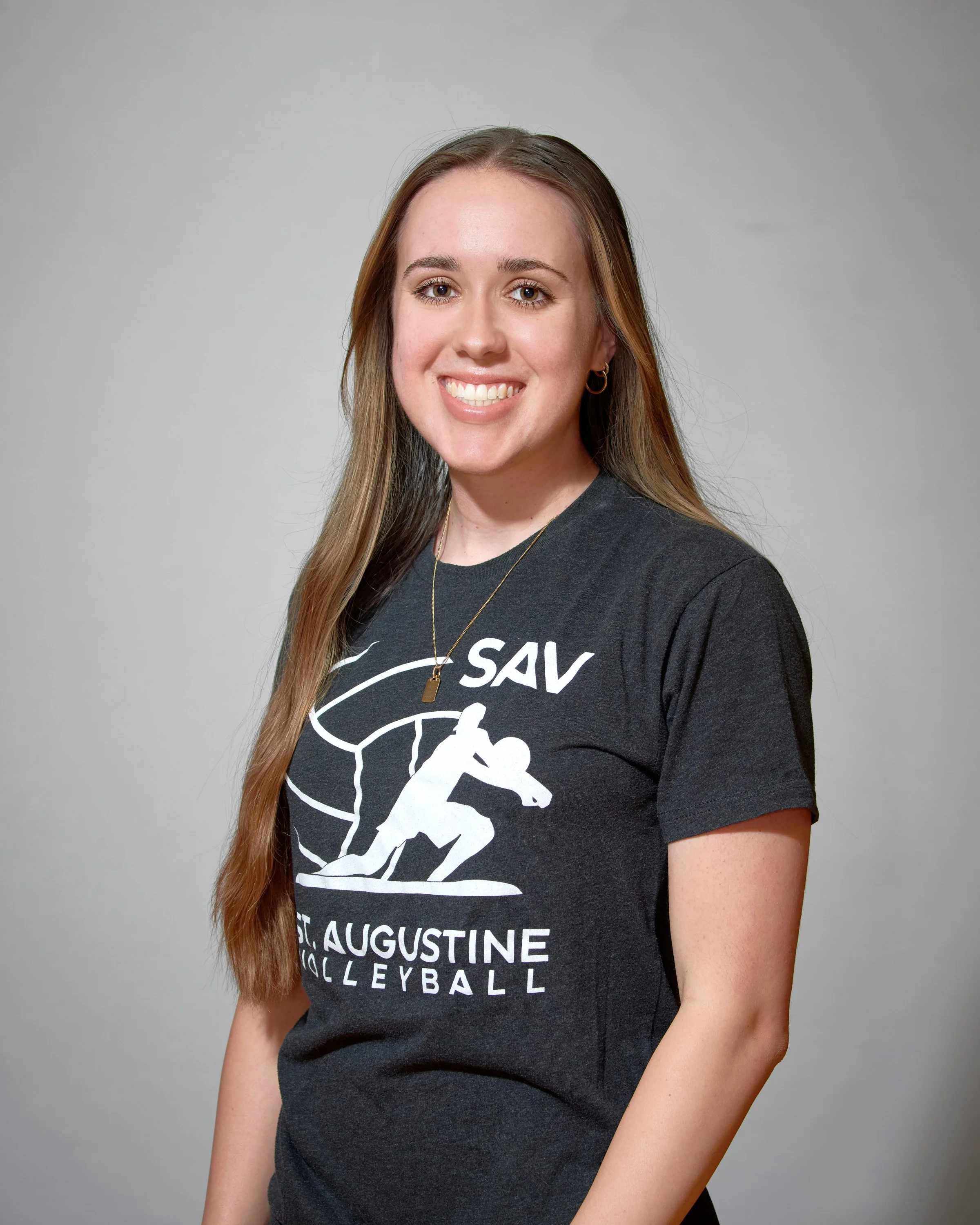 A young woman with long brown hair smiling, wearing a dark T-shirt with a volleyball graphic, a gold necklace, and hoop earrings.