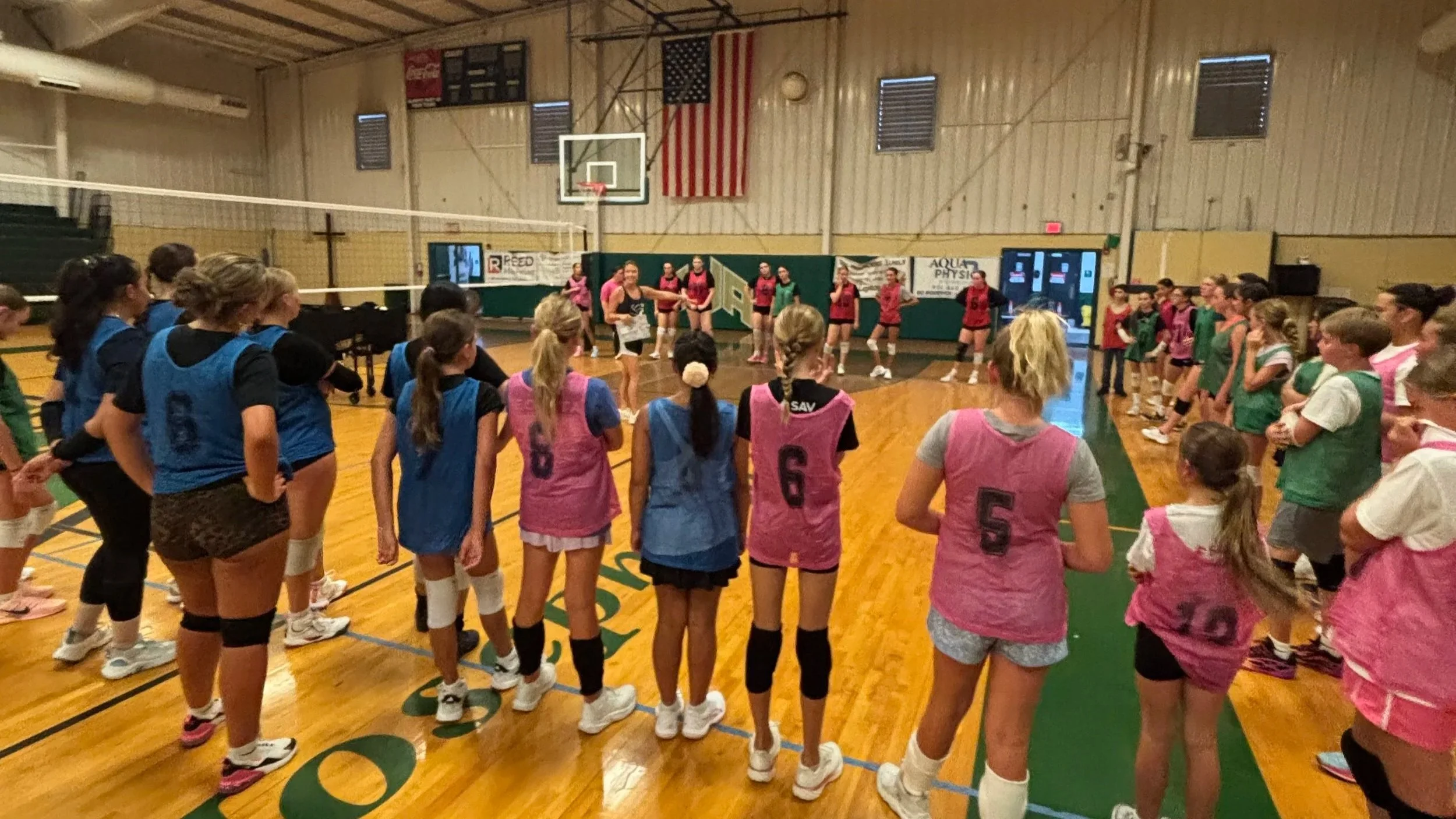 A group of young girls in pink and blue sports jerseys standing in a gymnasium, listening to a coach or instructor as they prepare for a volleyball game.