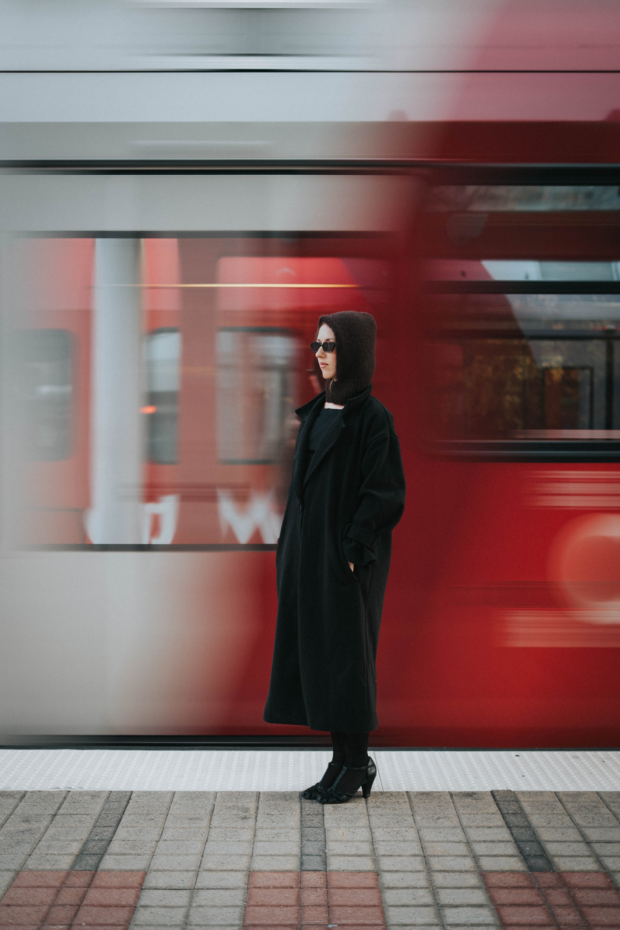A woman in a long black coat, black heeled shoes, sunglasses, and a hood stands on a city sidewalk in front of a red moving train at a station.