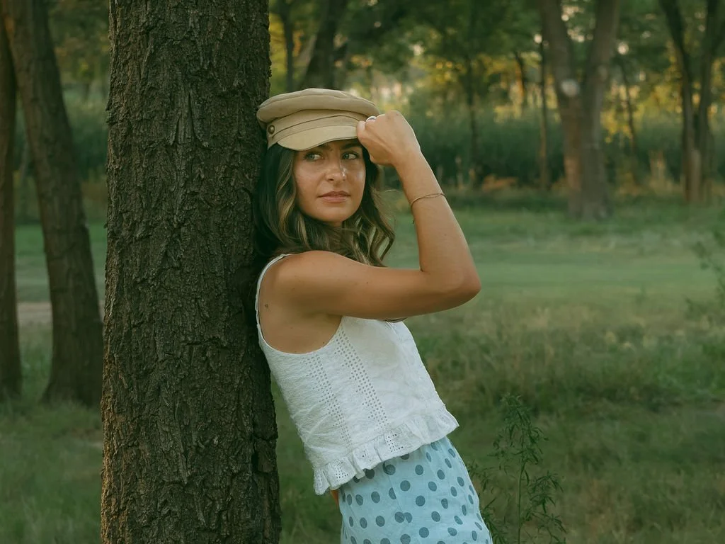 A woman leaning against a tree in a park or wooded area, wearing a beige cap, white sleeveless top, and blue polka-dot skirt, looking thoughtfully into the distance.