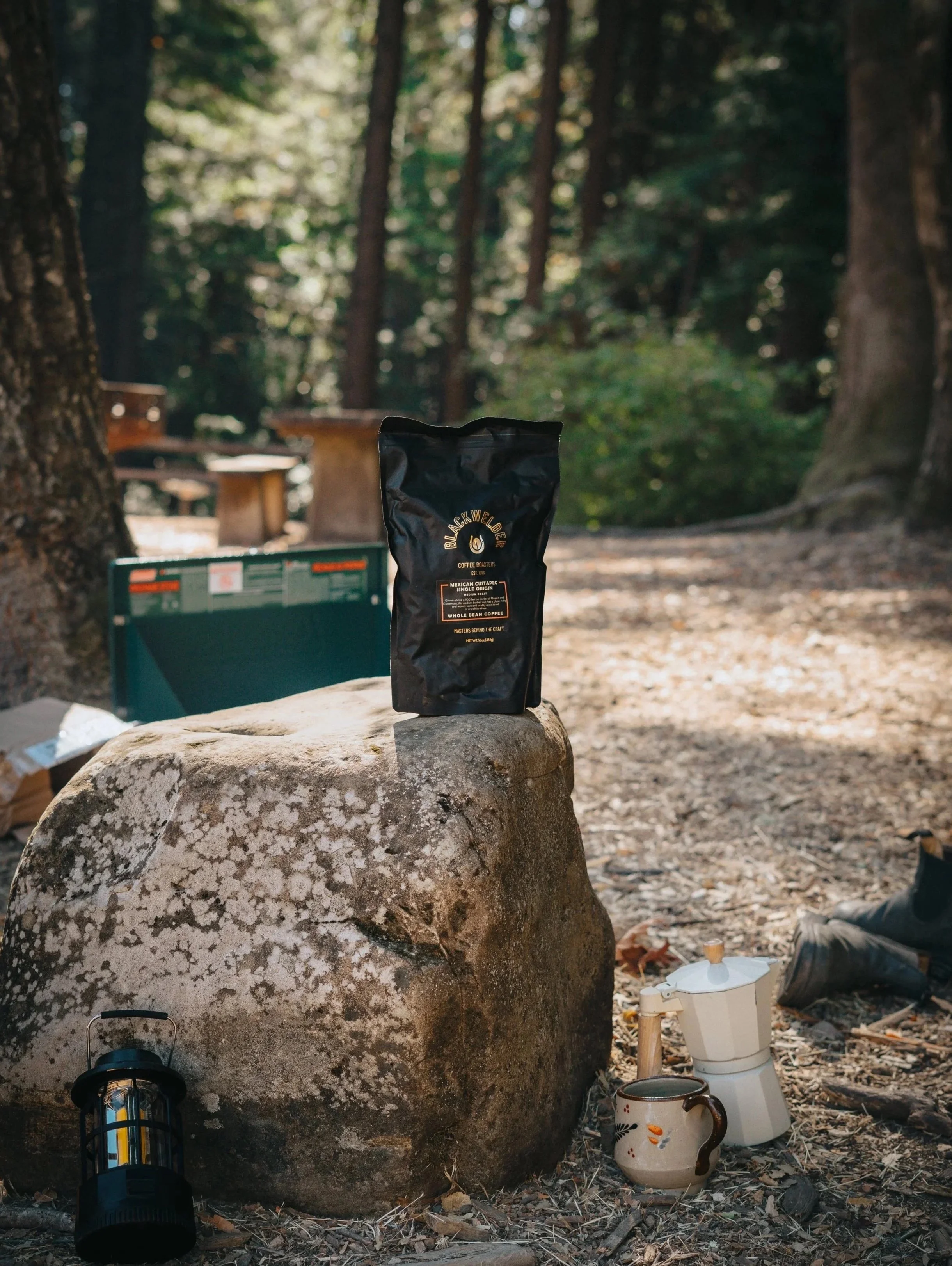 A bag of Blackwelder coffee roast placed on a large rock in a forest setting with trees and a wooden bench in the background, along with a lantern, mugs, and a small white lantern on the ground.