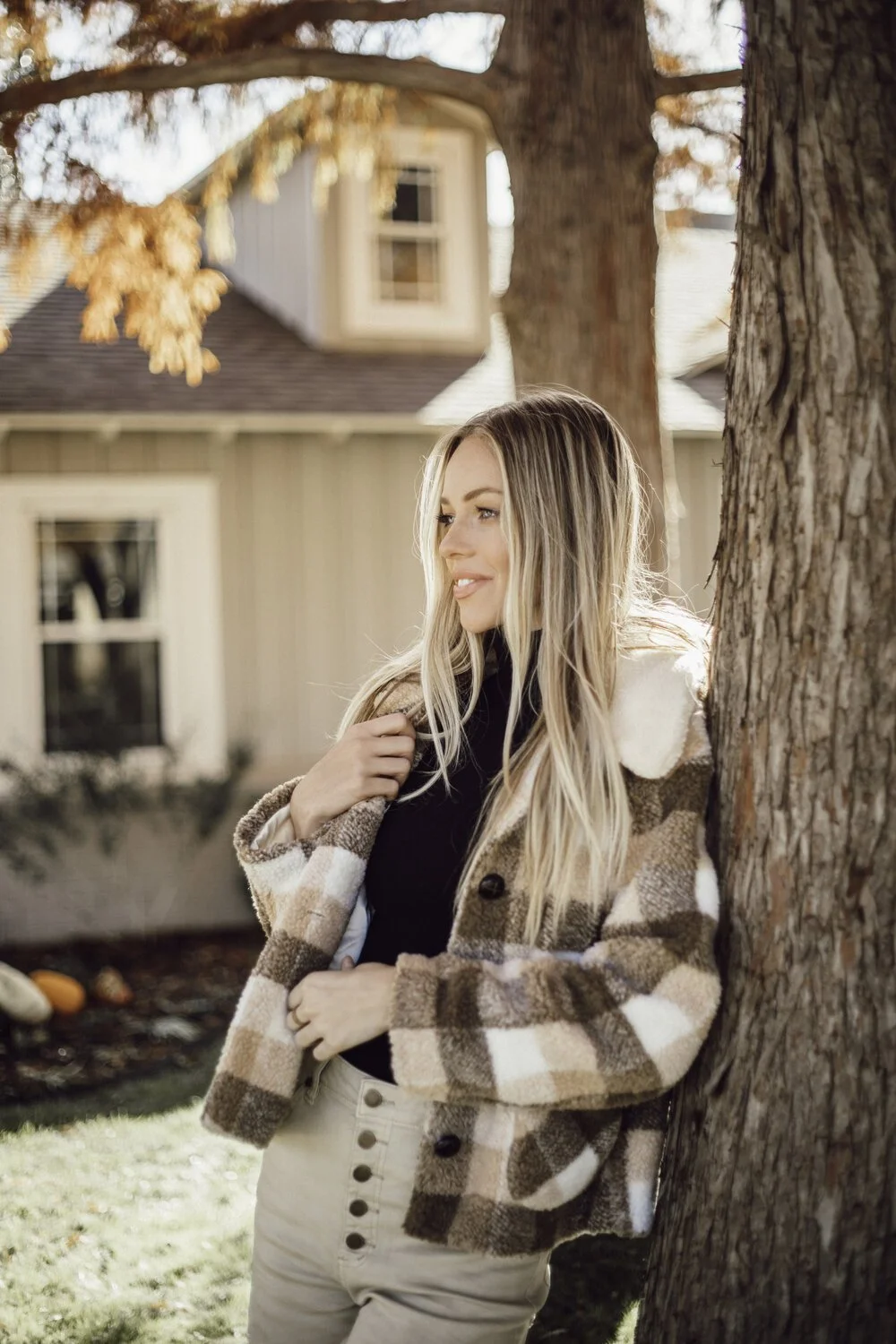 A young woman with long blonde hair leaning against a tree outdoors during autumn, wearing a checkered coat and beige pants, with a house and fallen leaves in the background.