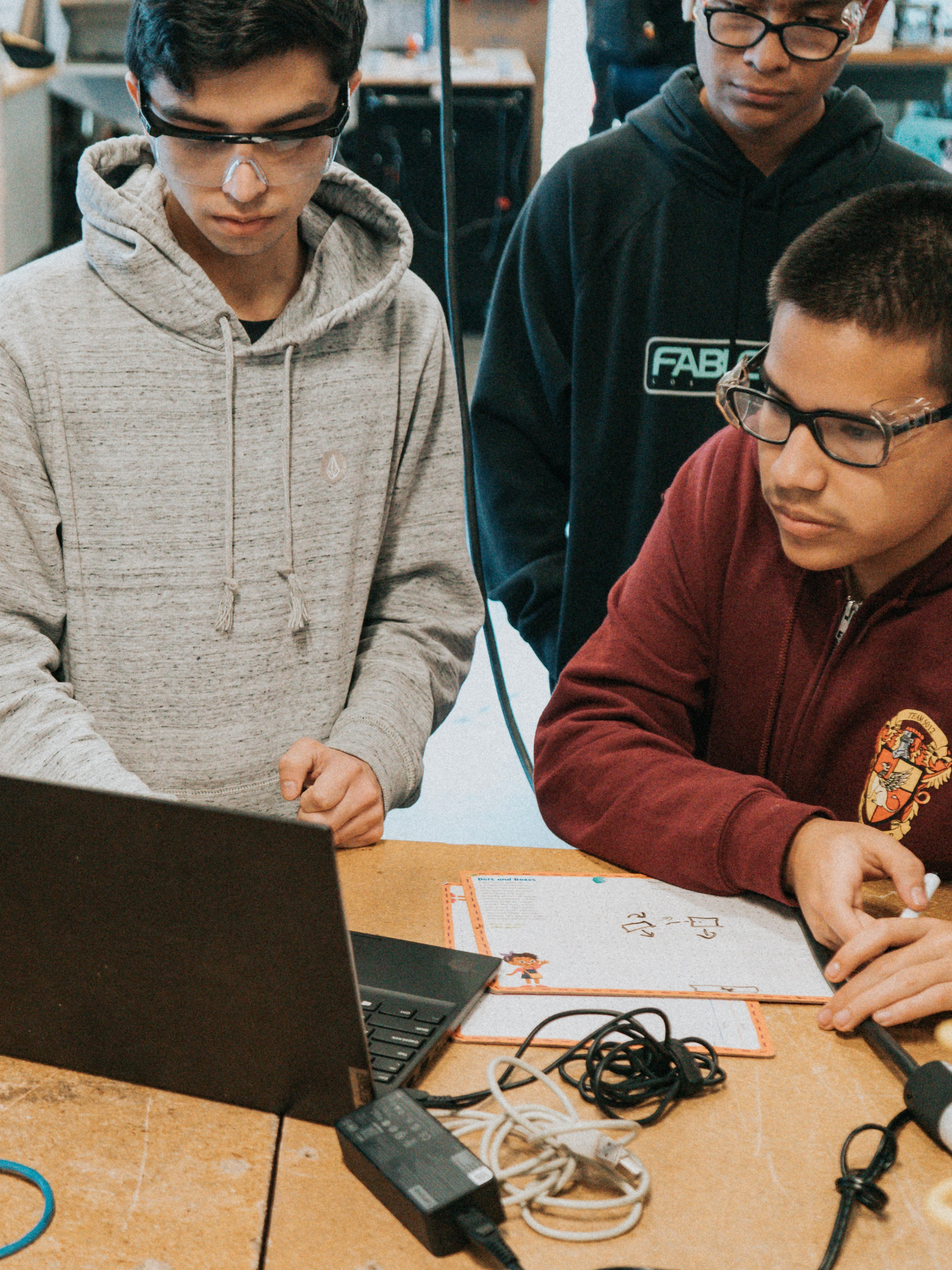 Three boys working together at a desk with laptops, cables, and papers in a classroom or lab setting.
