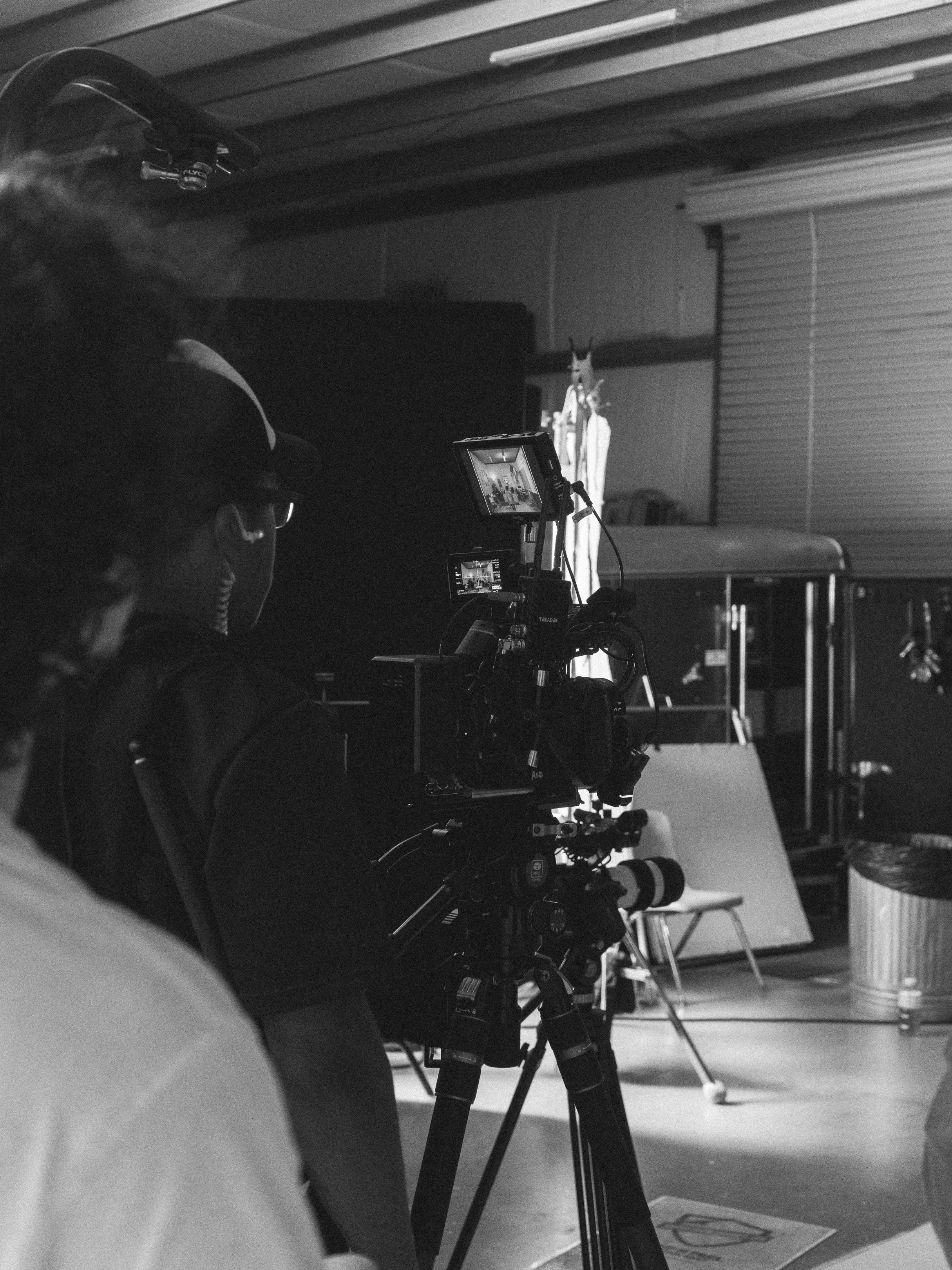 Black and white photo of a person operating a professional camera on a tripod in front of a backdrop, likely in a studio setting.