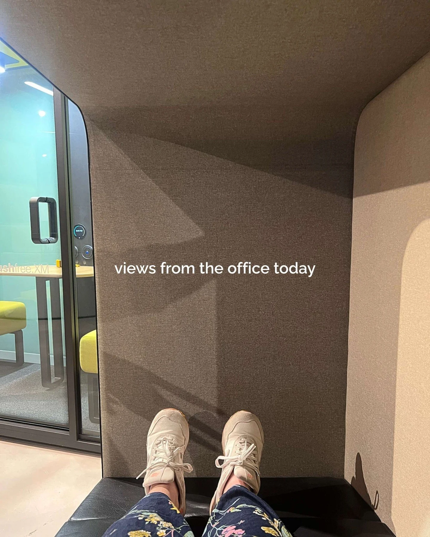Person wearing white sneakers and floral pants sitting in a chair in an office, with text on the wall that reads 'views from the office today' and a glass door to another room.