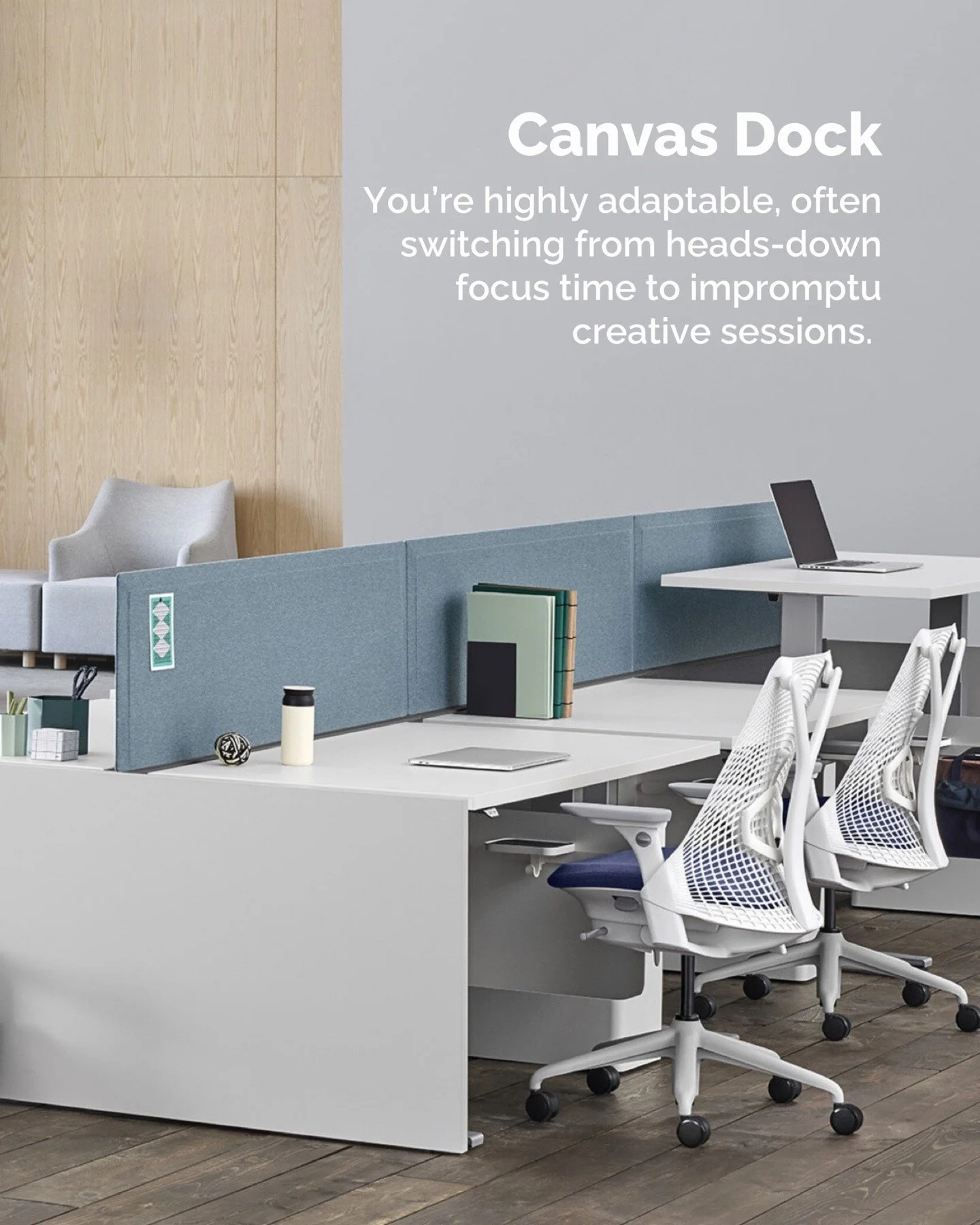 Modern office space with white desks, ergonomic mesh chairs, a cubicle with blue fabric dividers, and a laptop on one of the desks. There are items like books, a coffee cup, and a notebook on the desks. Background includes wooden wall panels and a large gray wall with text about the workspace.