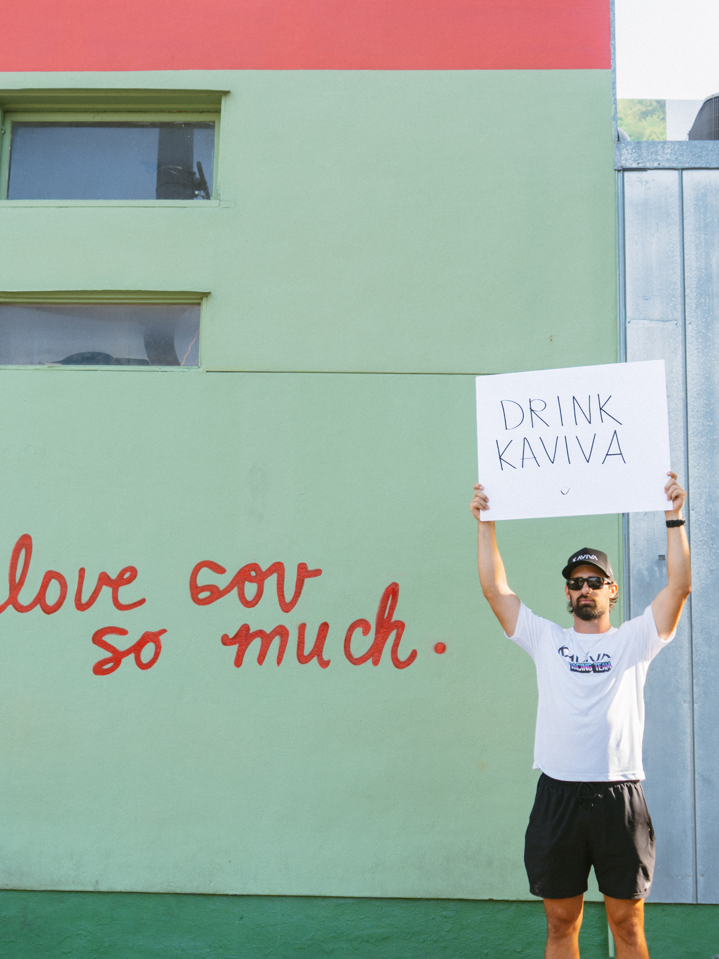 A man wearing sunglasses, a white t-shirt with the word 'KAVIVA' and additional text, and black shorts, holding a white sign that says 'DRINK KAVIVA' in front of a green wall with red handwritten text.