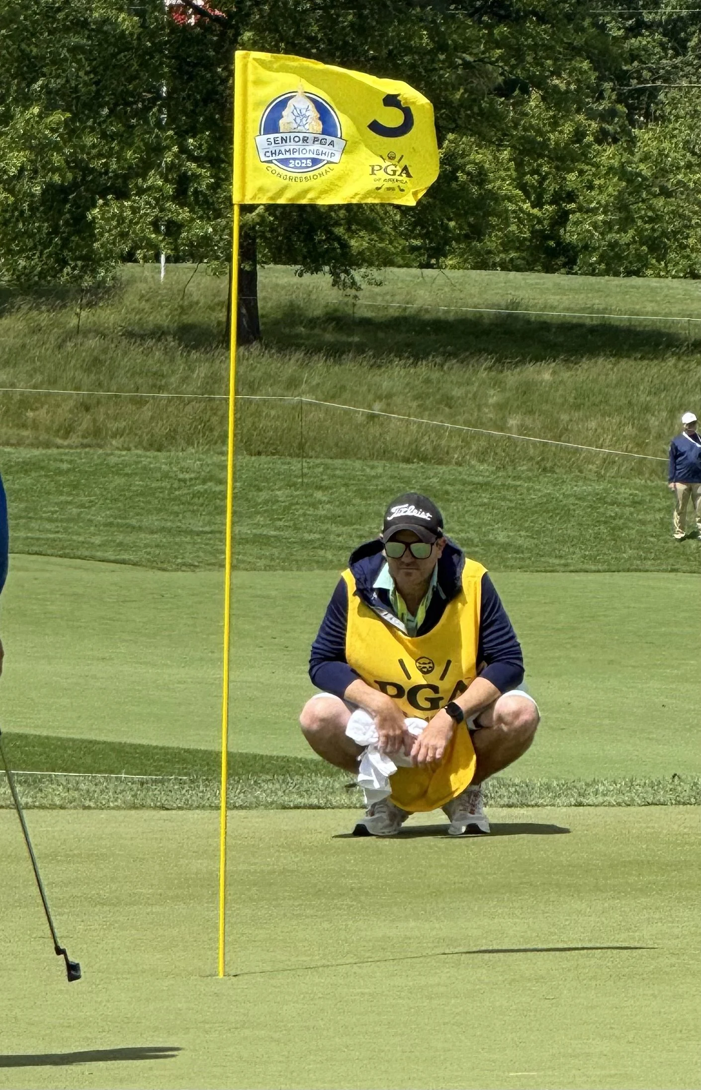 A golfer crouching on a golf course near the hole with a yellow flag marked with the number 5 and PGA sponsorship logo, during a senior championship in 2023.