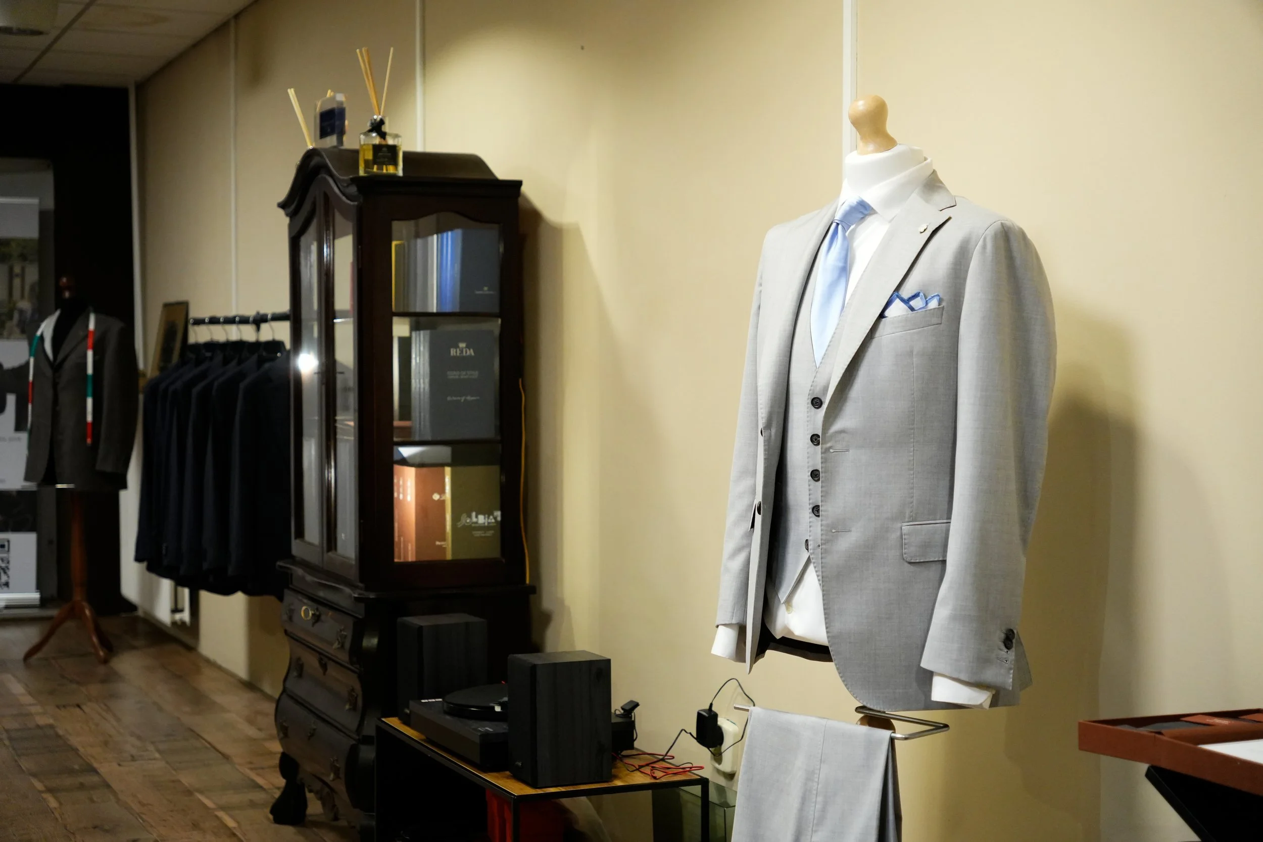 Een mannelijke mannequin gekleed met een lichtgrijze pak en een blauwe das, in een winkel met kleding en accessoires.