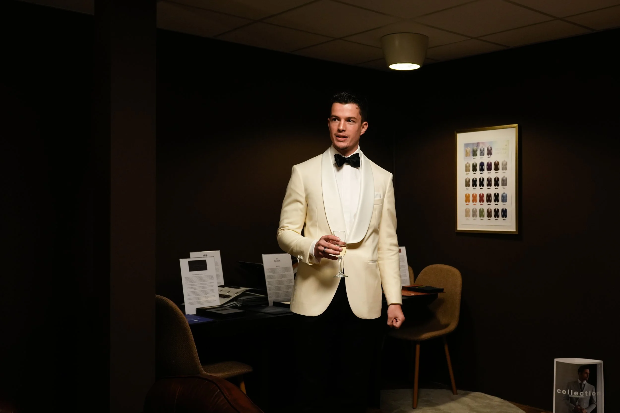 Man in een witte tuxedo met zwarte strik houdt een glas wijn en staat in een donkere ruimte met kunst aan de muur.