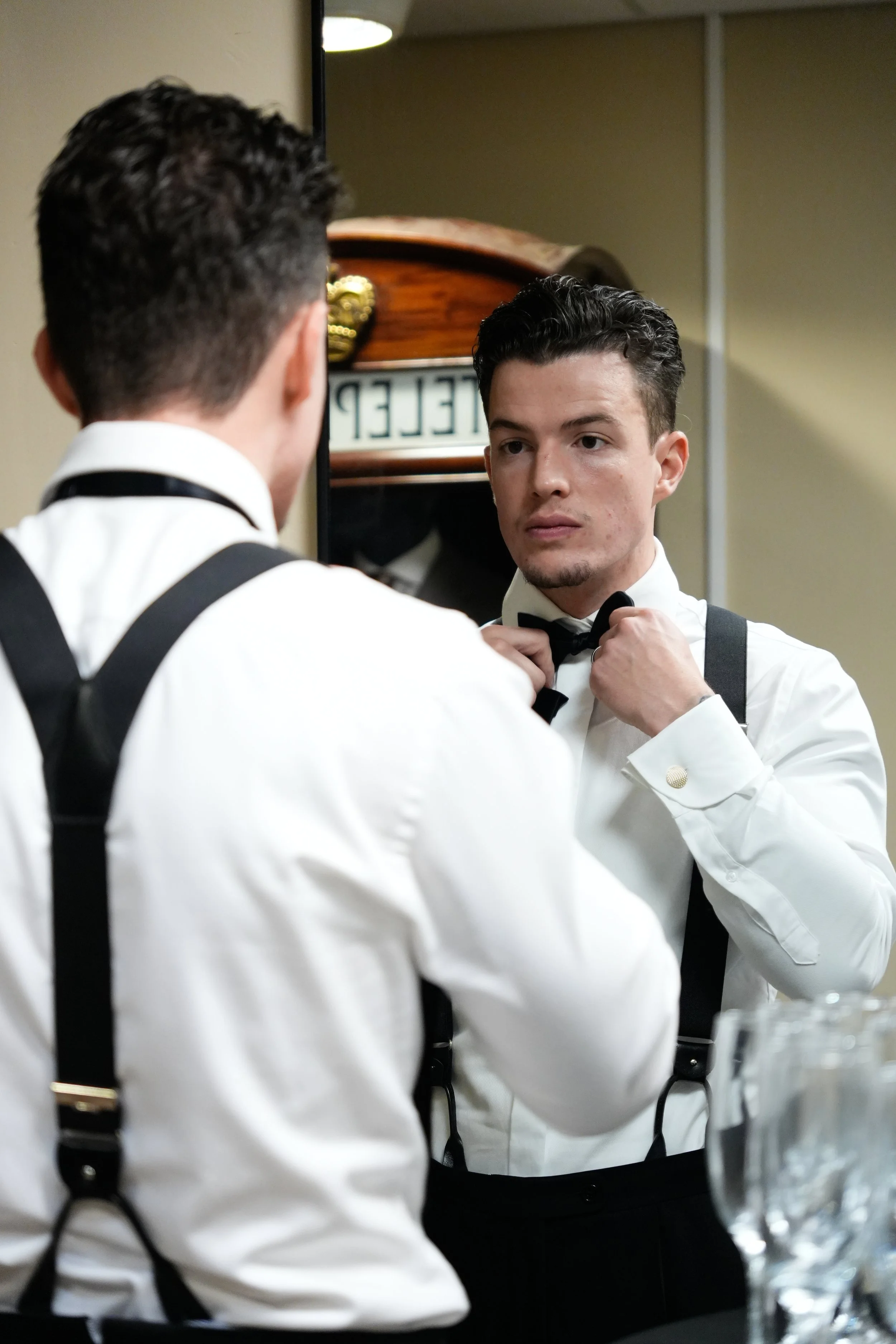 Een man in witte overhemd en zwarte bretels die zijn tuniek rechtmaakt terwijl hij in de spiegel kijkt.