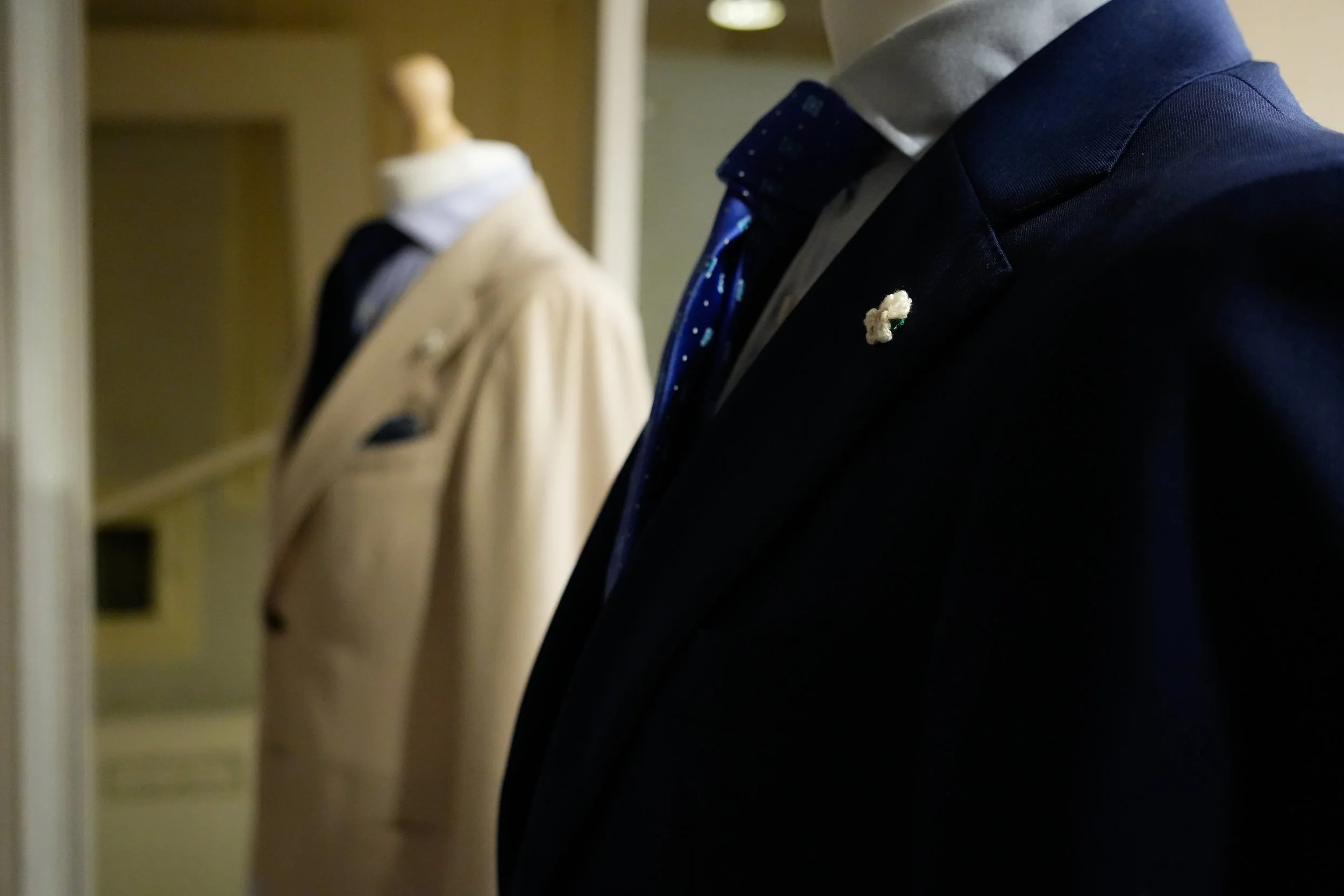 Close-up van een man in een donkerblauw pak met een donkerblauwe stropdas met witte stippen, en een wit overhemd, met een kleine bloem als broche op de revers.