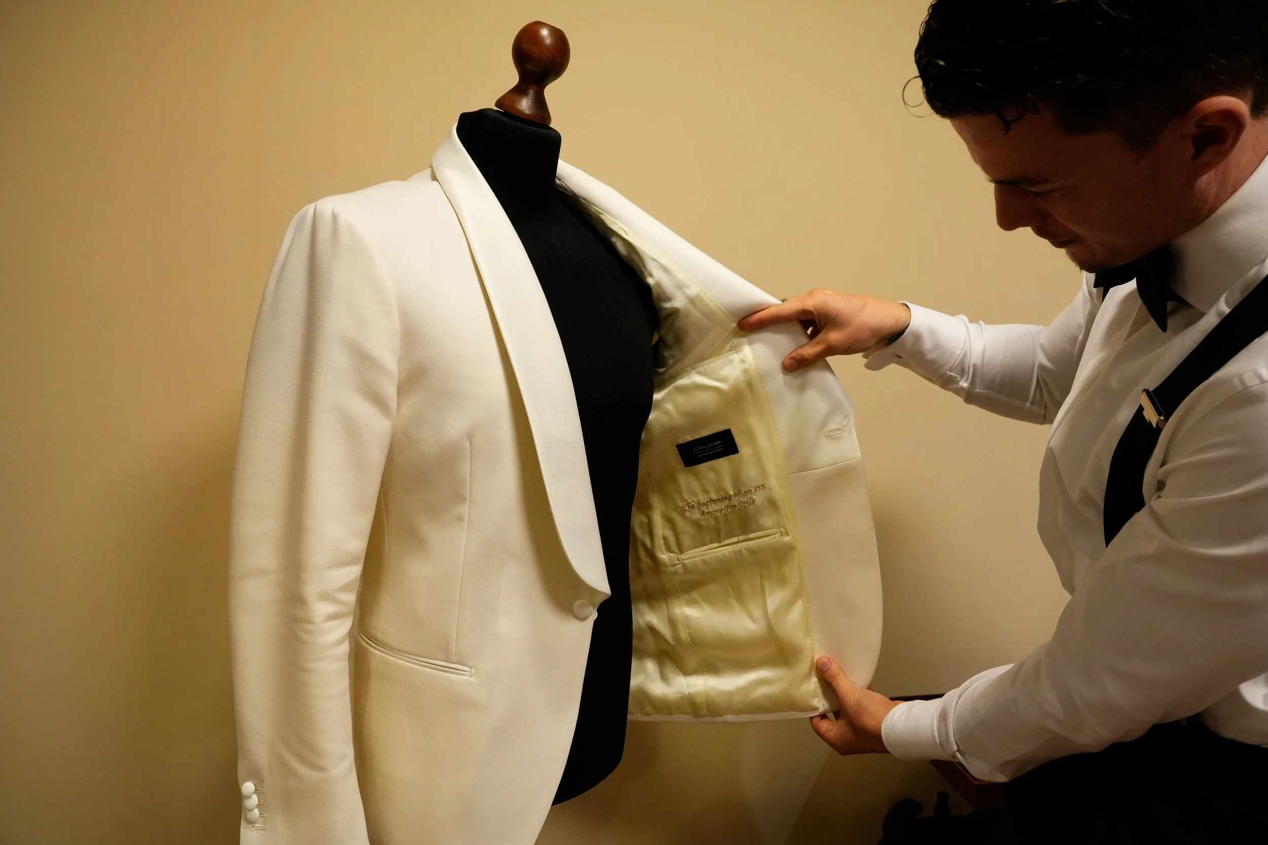 Man opent een luxe witte smokingjas op een kledingmal, tegen een beige muur.