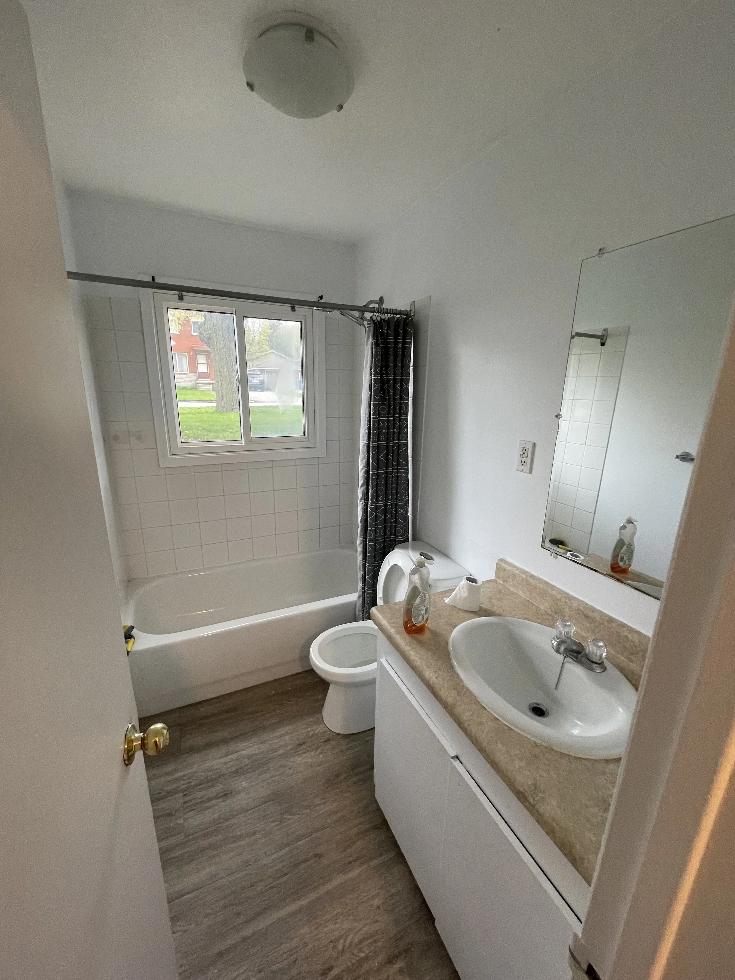 A small bathroom with a bathtub, a window with a view of a yard, a toilet, a sink with a mirror, and cleaning supplies on the counter.