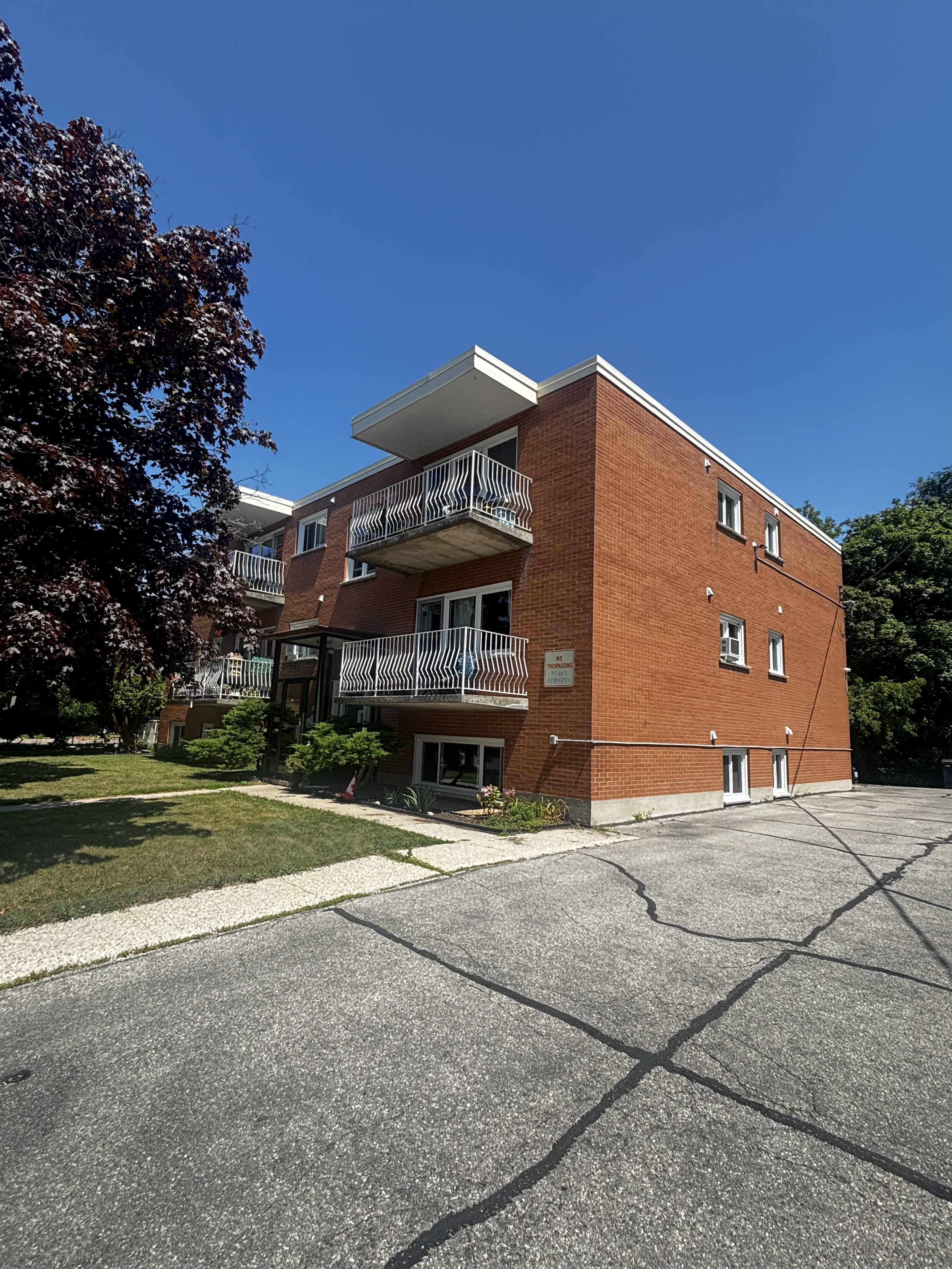 A three-story brick apartment building with trees in the front yard, a sidewalk, and parked cars on the street.