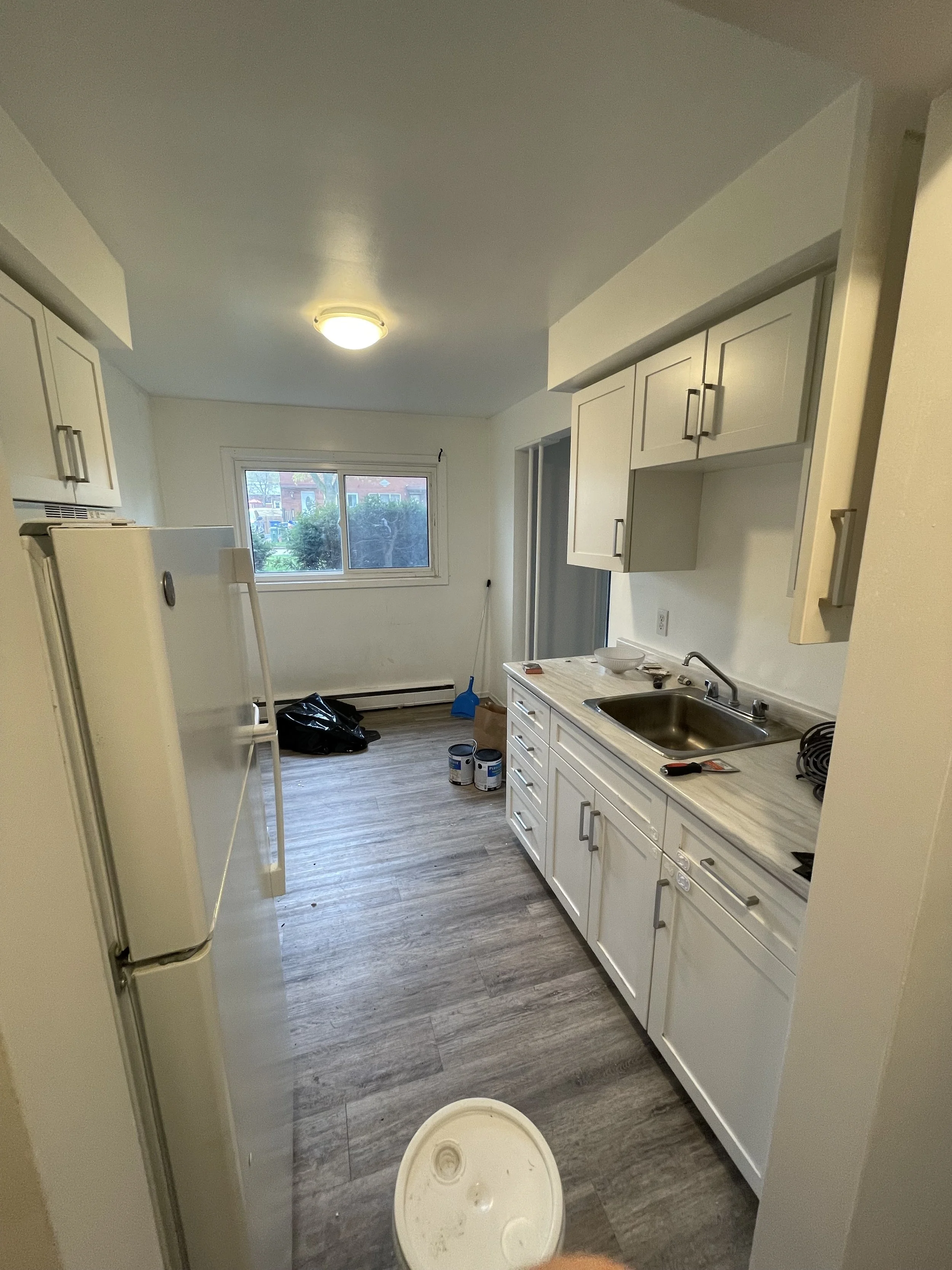 Empty kitchen with white cabinets, a stainless steel sink, a window with a view of outdoors, and cleaning supplies on the floor.