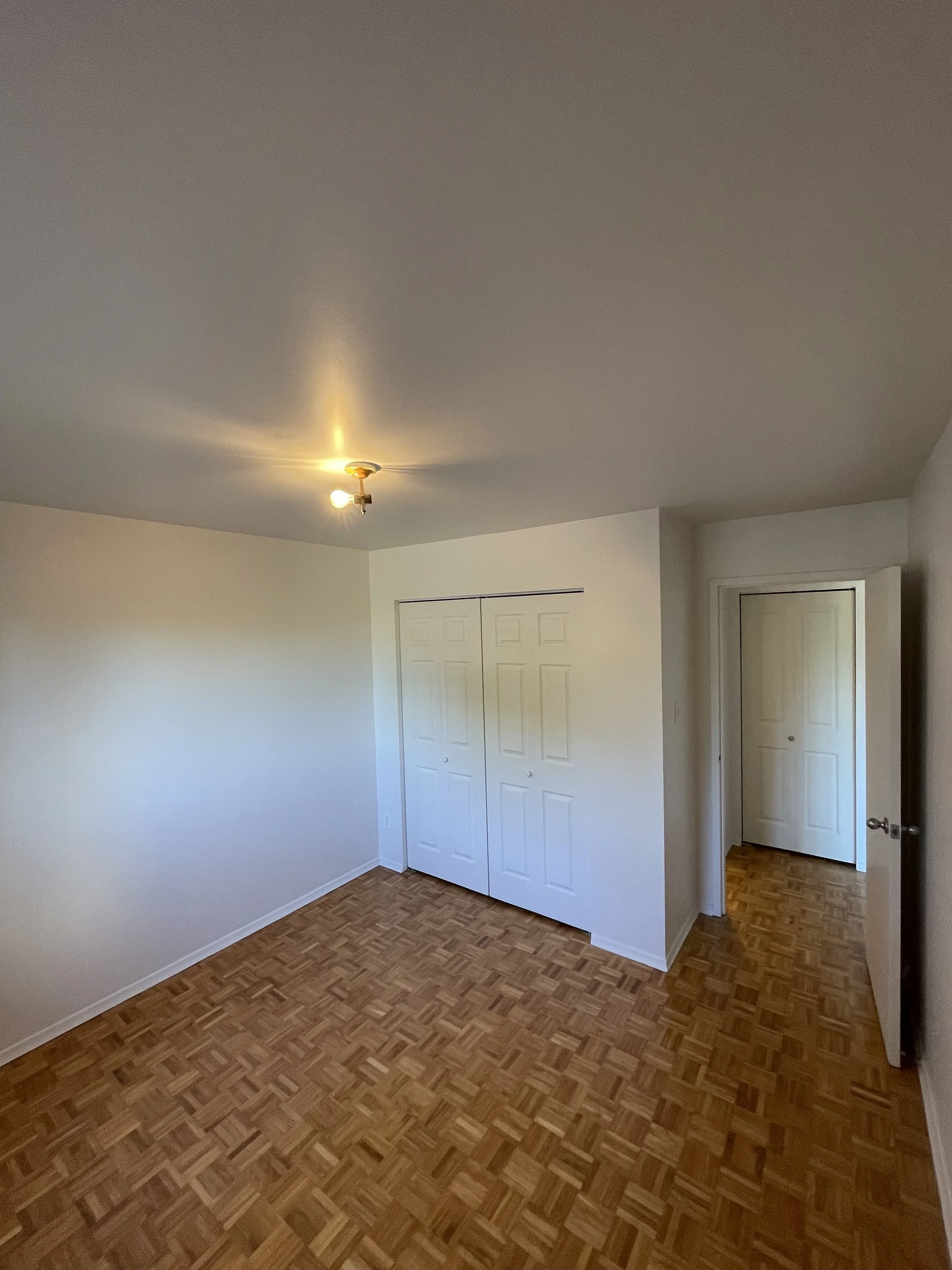 Empty bedroom with white walls, parquet flooring, a ceiling light fixture, closed closet doors, and an open door leading to a hallway.
