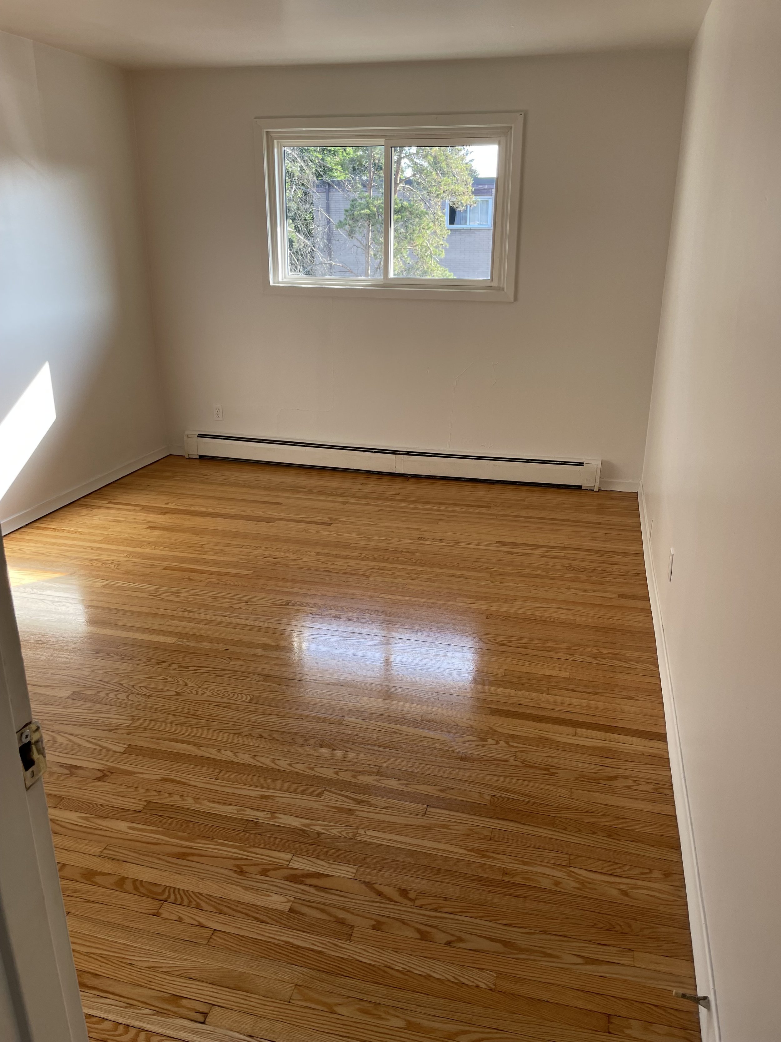 Empty room with hardwood floors, white walls, a window showing trees and a building outside, and a baseboard heater along the wall.