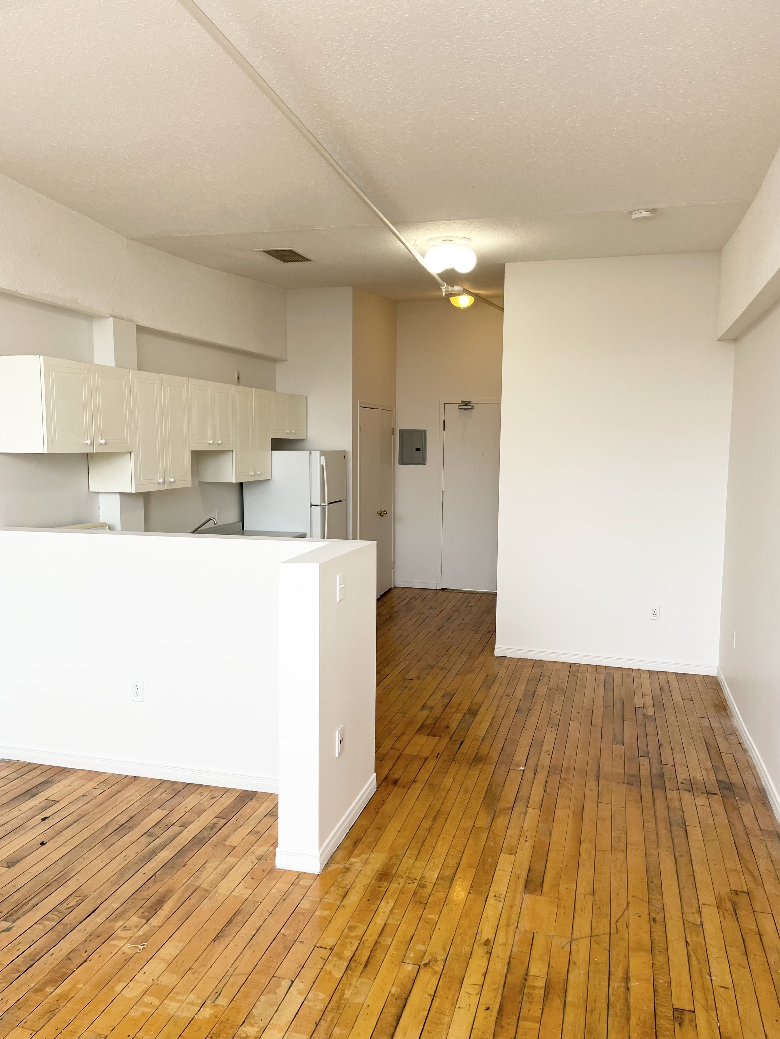 An empty apartment with hardwood floors, a kitchen with white cabinets and a refrigerator, and a small entryway area with a door and an electrical panel.