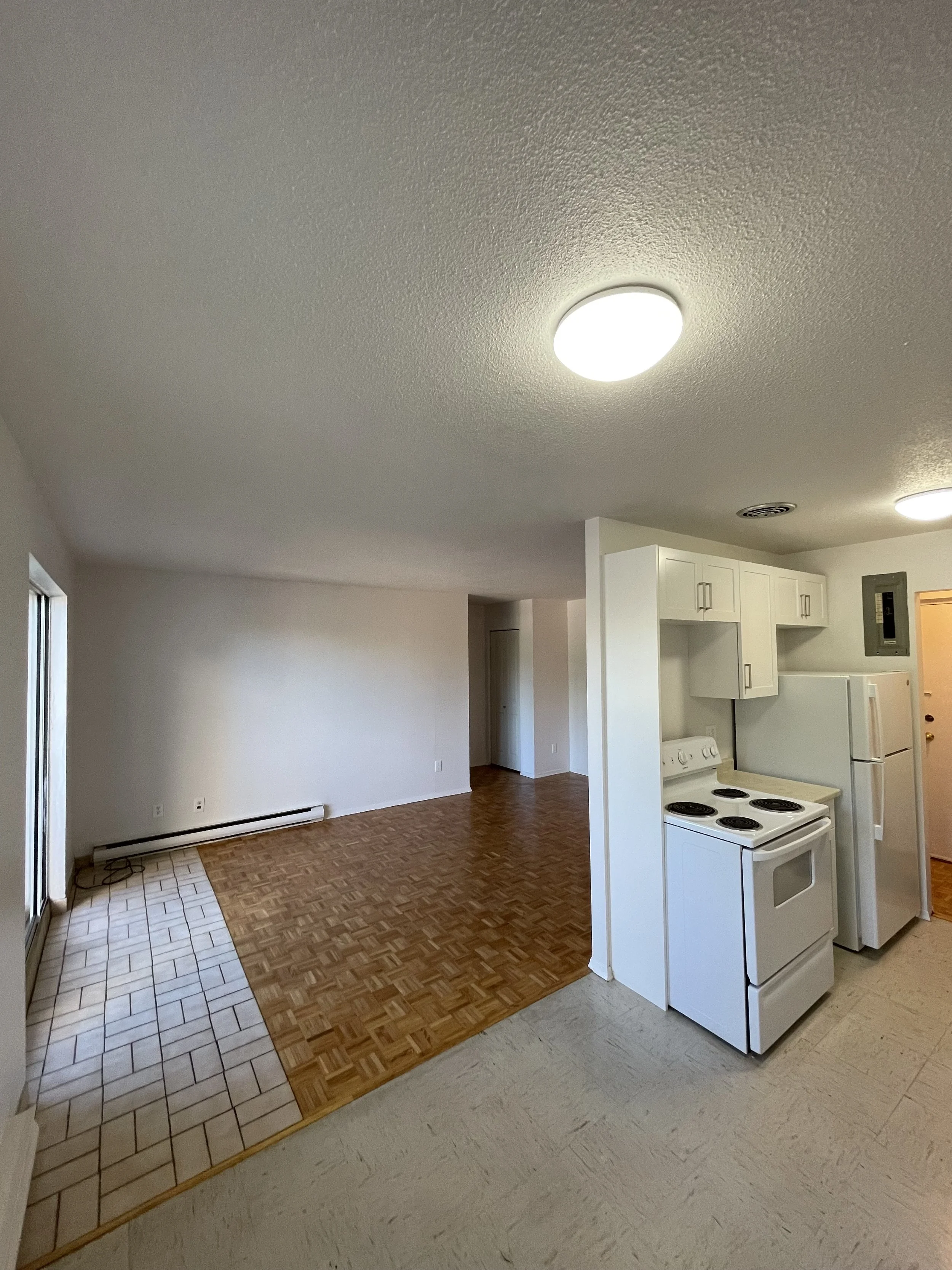 Empty living room and kitchen area with white cabinets, white stove, and refrigerator, featuring a mix of tiled and wood flooring.