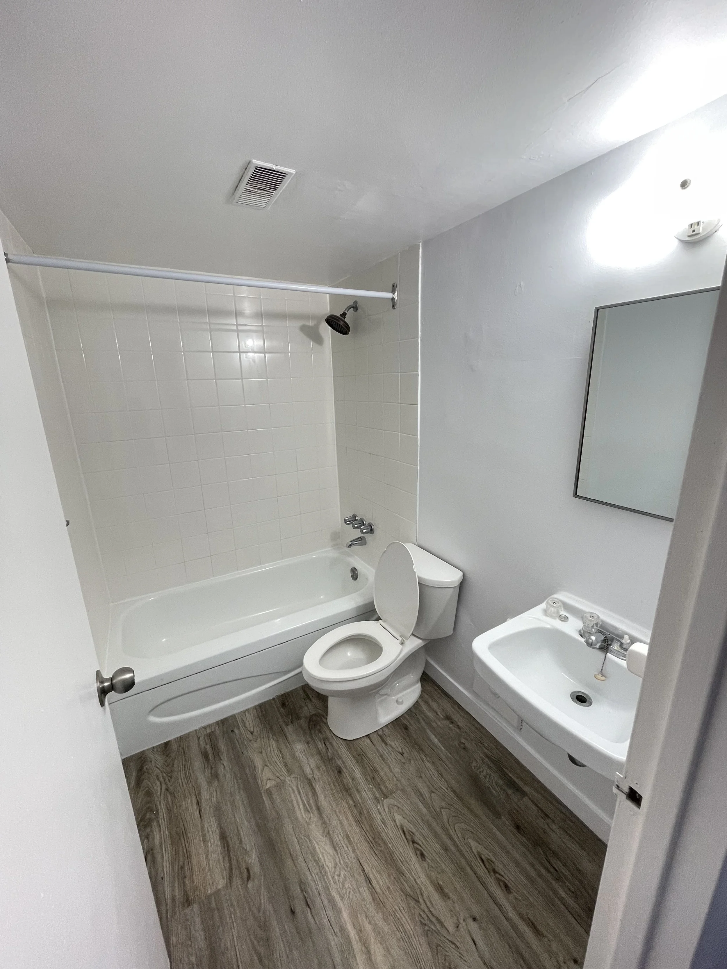 Small bathroom with a bathtub, toilet, and small sink with a mirror above it. The floor is wood-like vinyl, and the walls are plain white. There is a shower curtain rod over the bathtub.