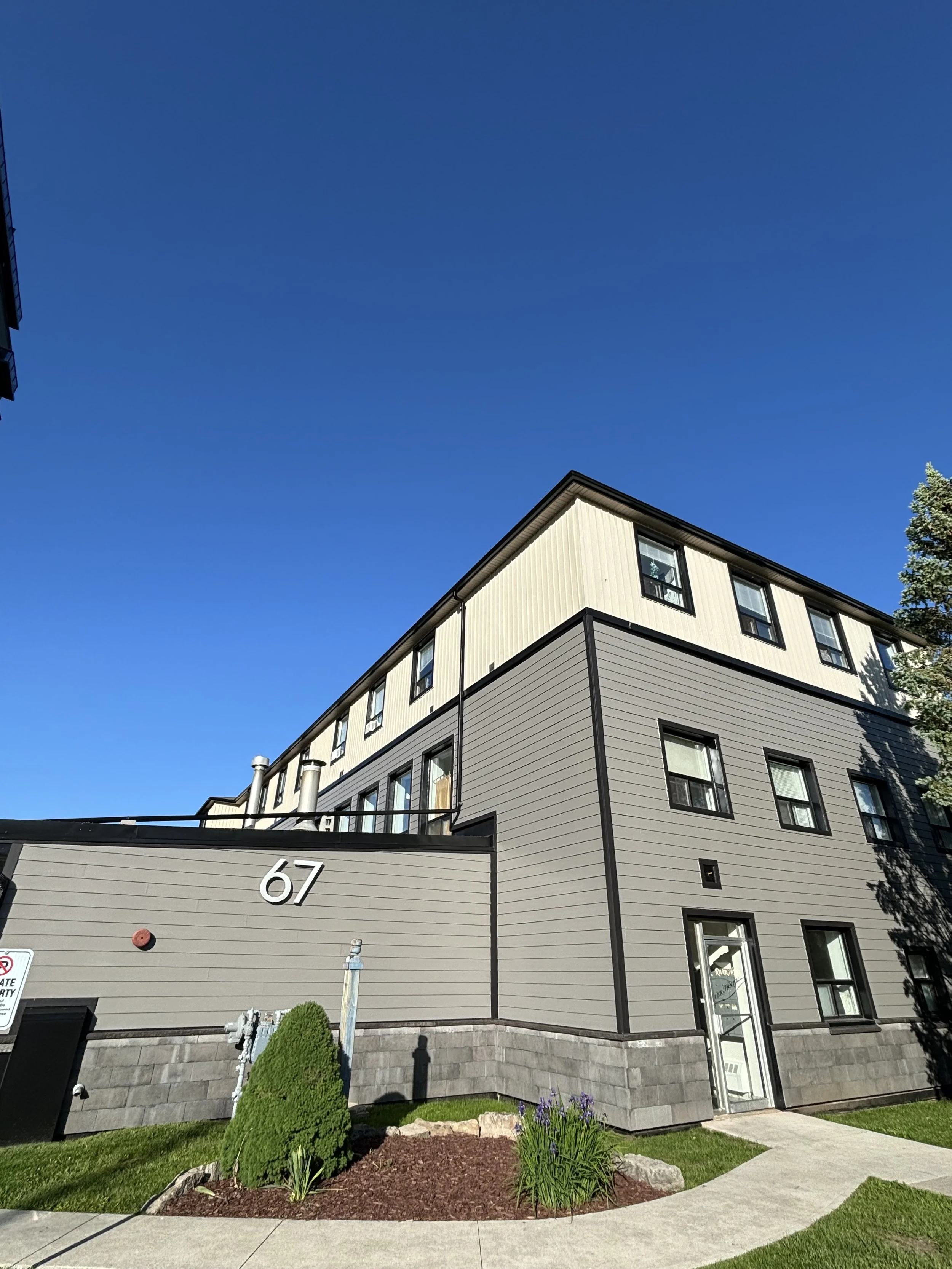Multiple-story residential building with beige and gray siding, black-framed windows, and a glass door at the entrance, numbered 67, under a clear blue sky, with landscaped grass, shrubs, and a sidewalk in the foreground.