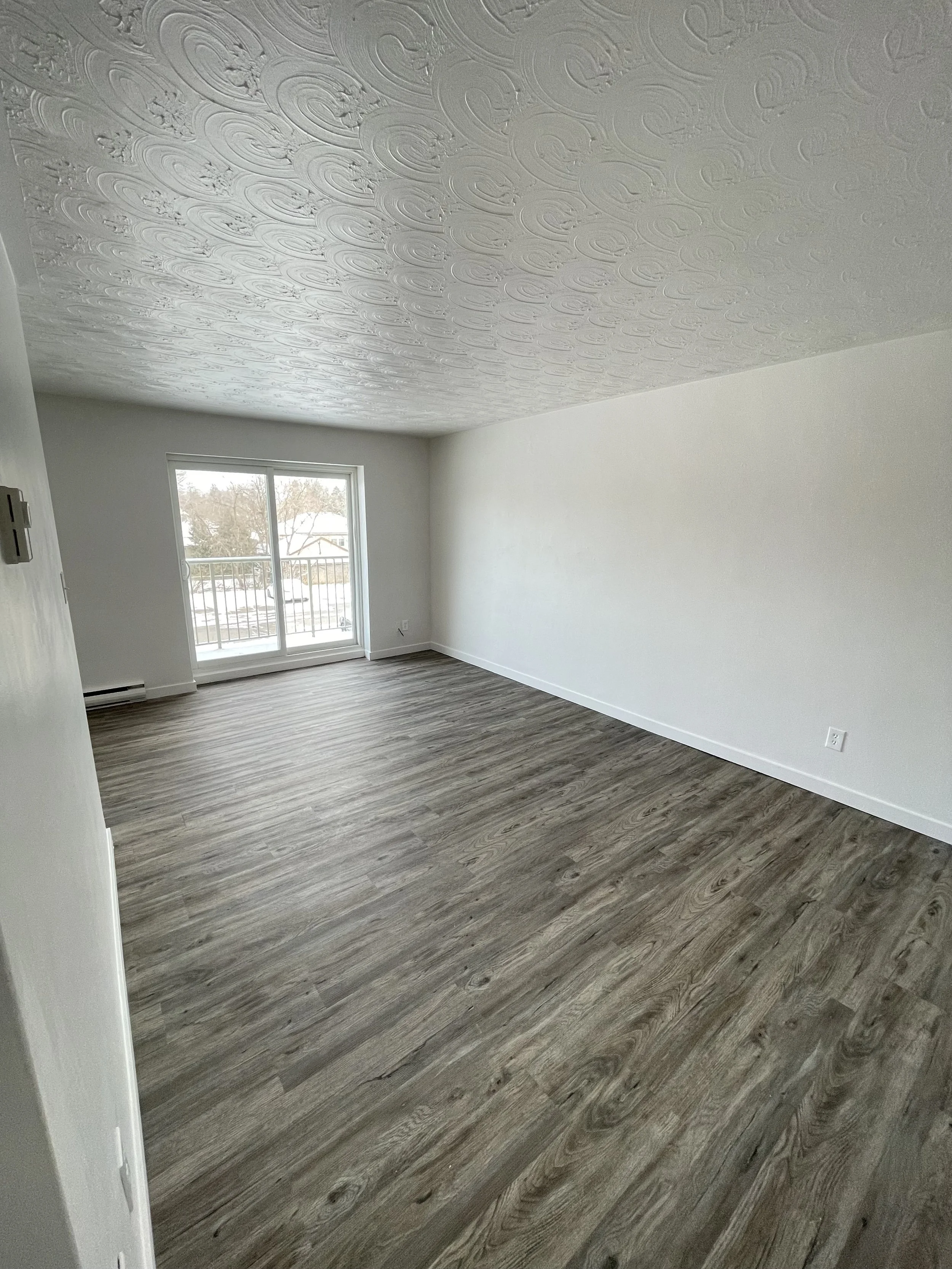 Empty living room with wood laminate flooring, white walls, textured ceiling, and sliding glass door leading to a balcony with a view of trees and snow.