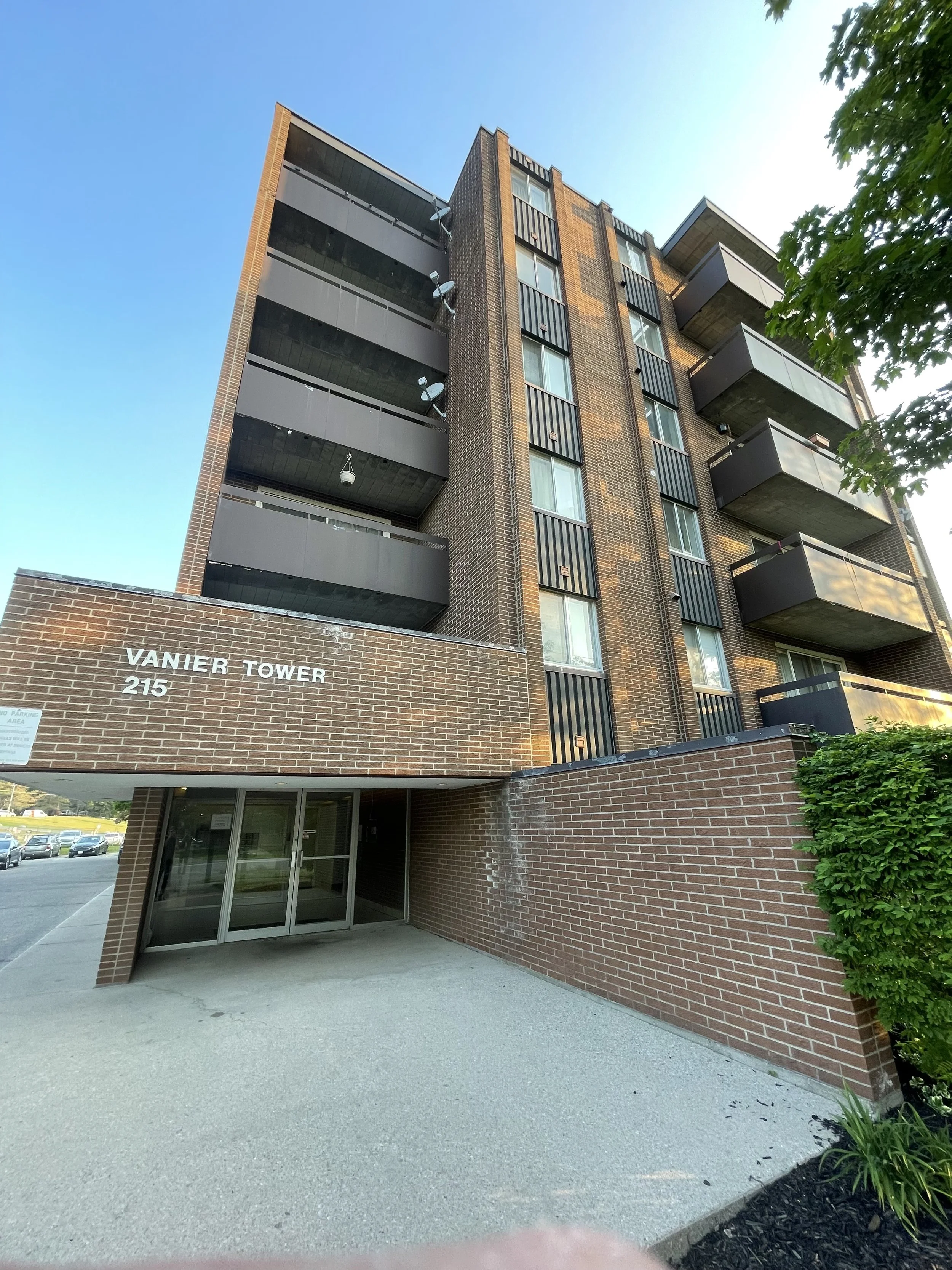 A multi-story brick apartment building named Vanier Tower, number 215, with balconies and a glass entrance door at the ground level, surrounded by trees and parked cars.