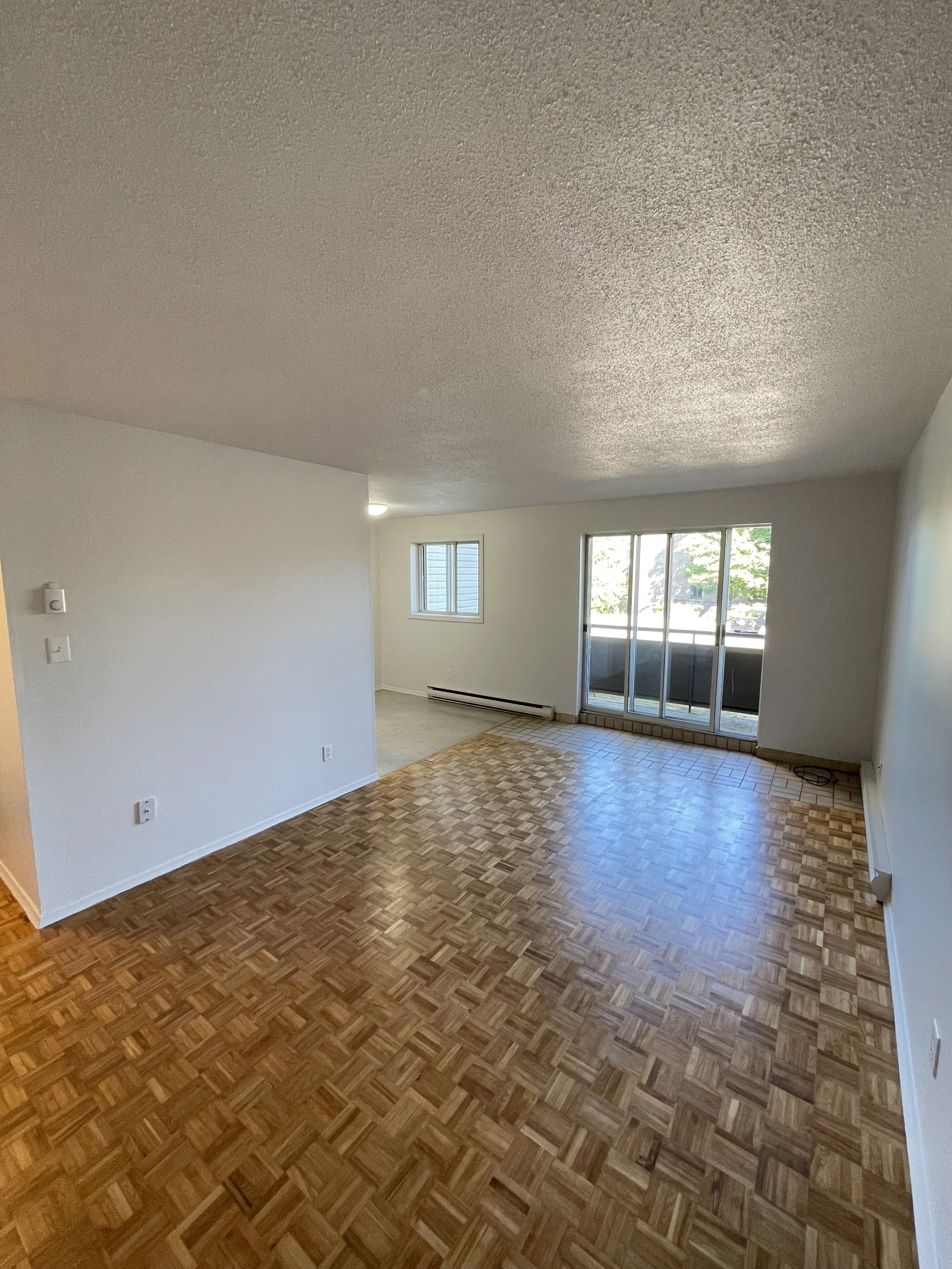 Empty living room with wood parquet flooring, white walls, a large sliding glass door, and a window, with a small balcony visible outside.