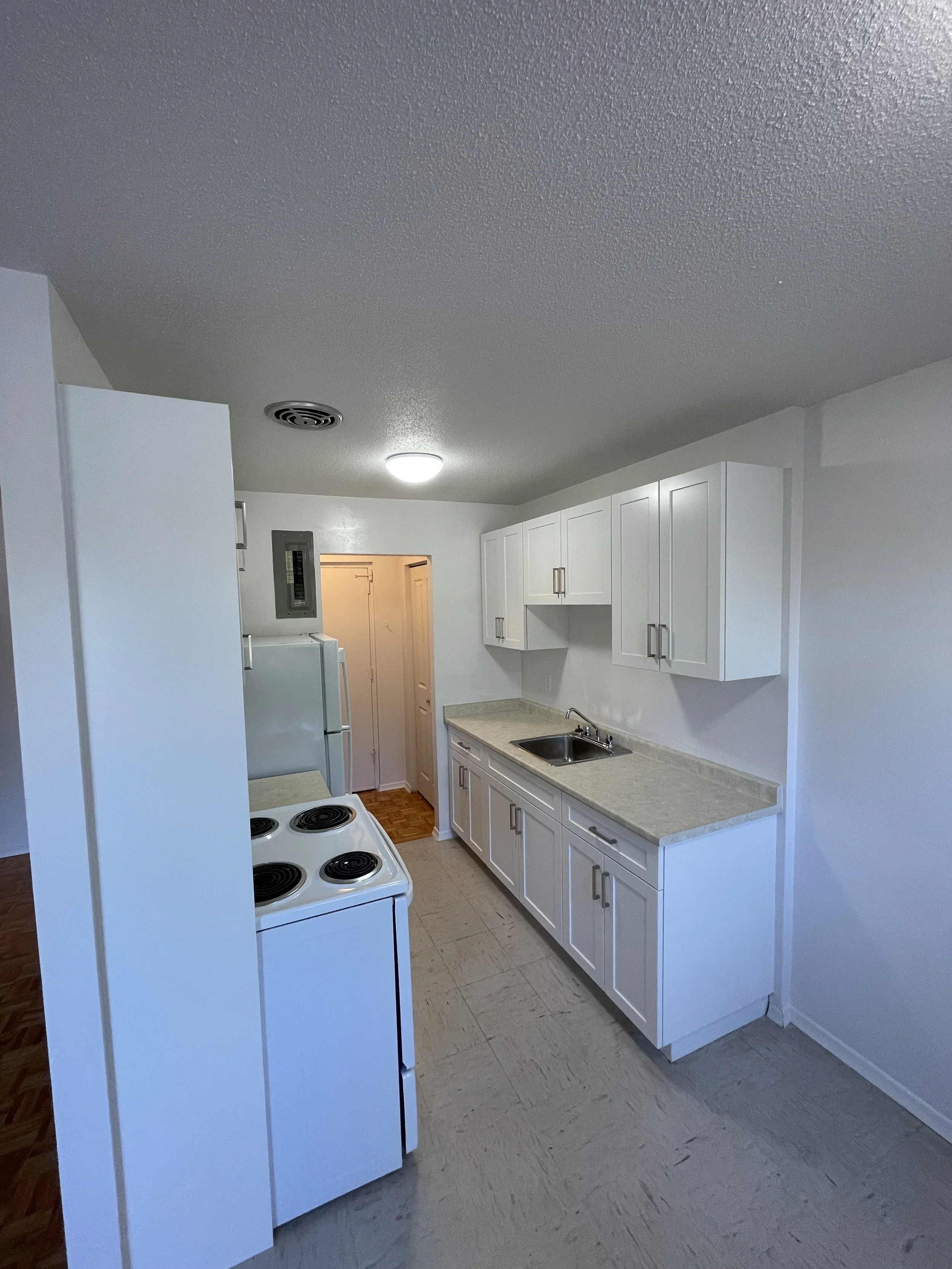 Kitchen with white cabinets, beige countertops, a stainless steel sink, a white stove, and a white refrigerator. The kitchen is lit by ceiling lights, with carpeted flooring and a textured ceiling.