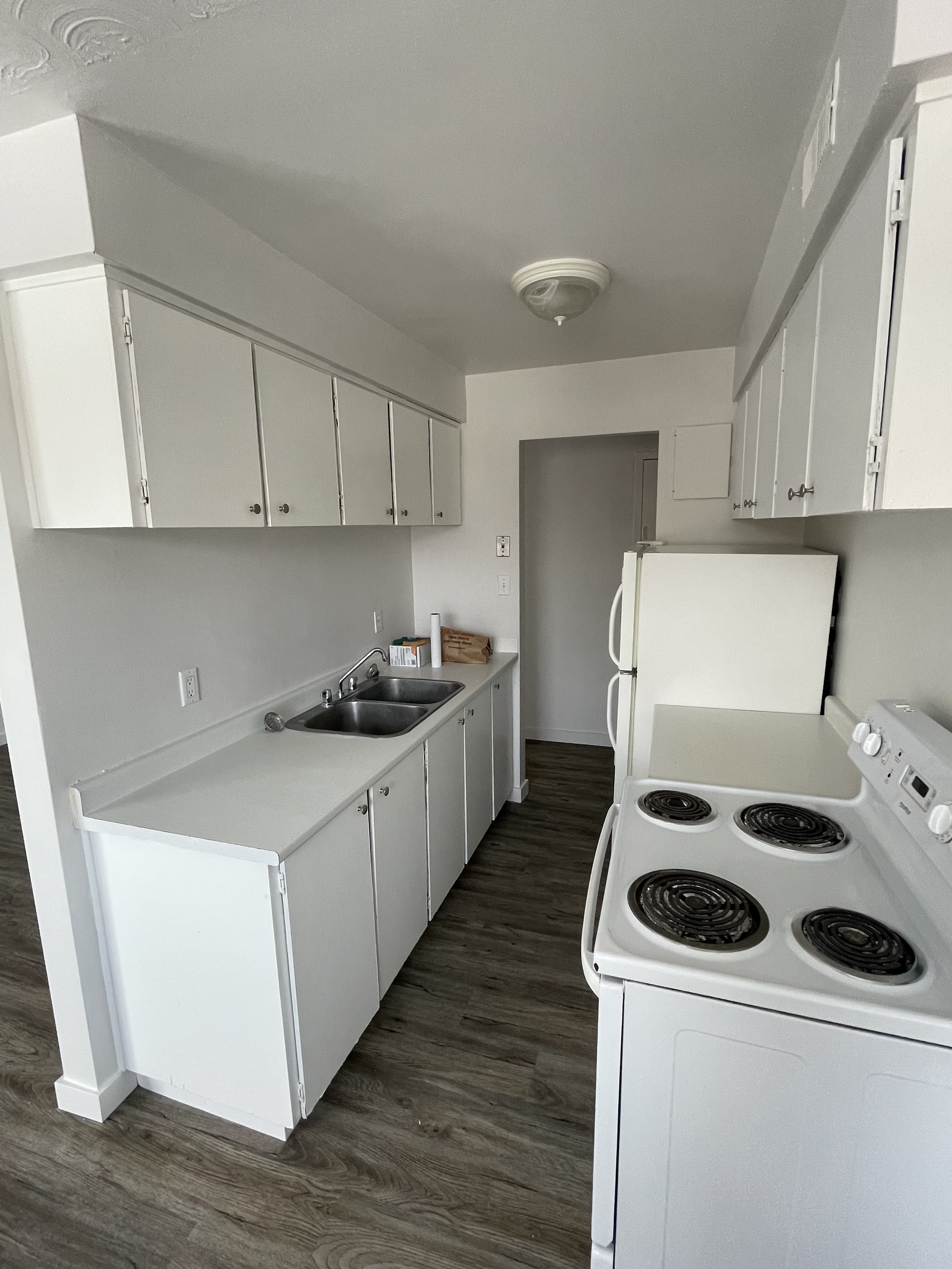 Empty kitchen with white cabinets, a double sink, a white electric stovetop, and a refrigerator against the wall. The floor is wood-like laminate, and the walls are painted white.