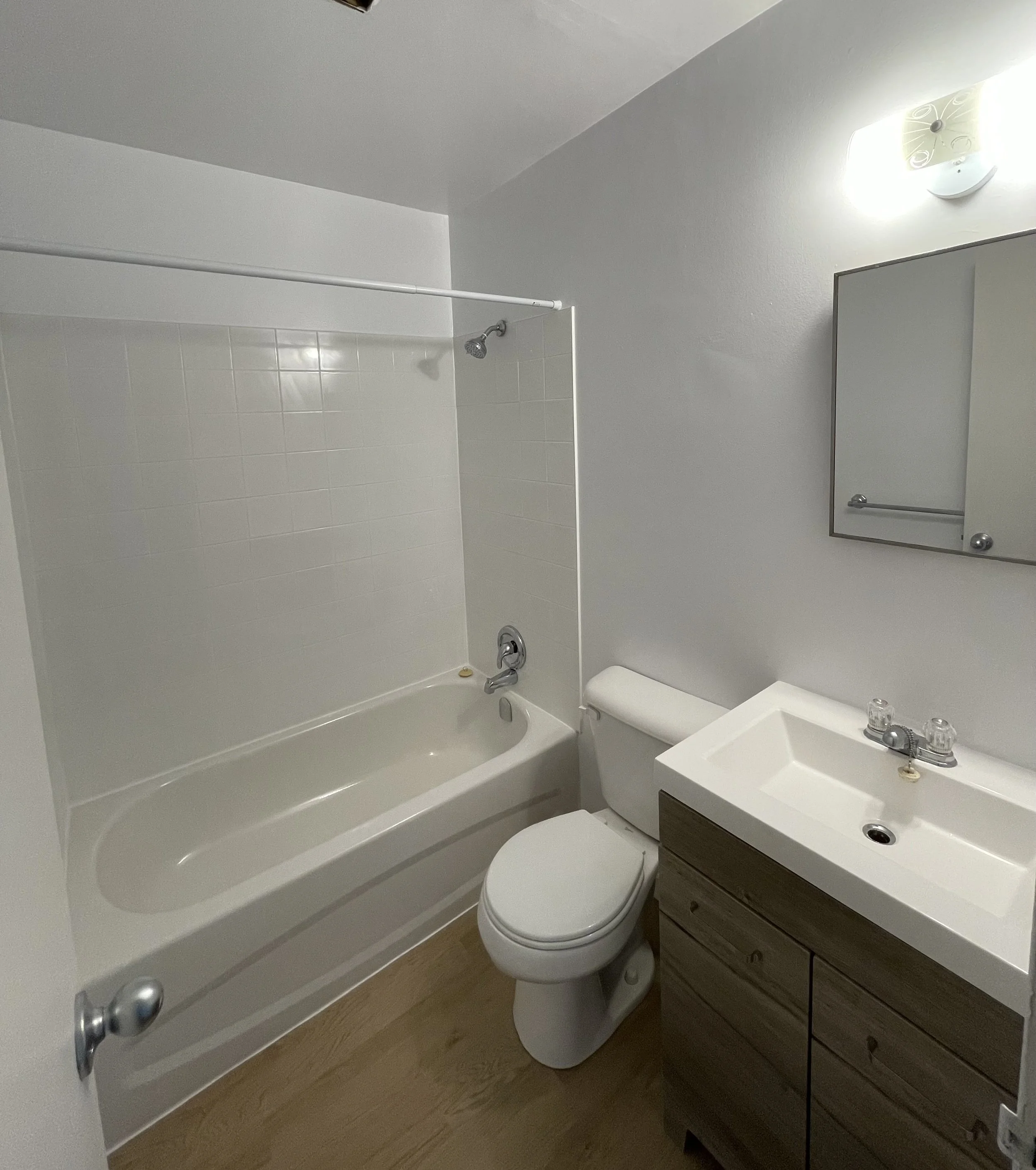 A small bathroom with a bathtub and shower on the left, a toilet in the middle, and a small sink with a cabinet on the right, with a mirror and a light above.