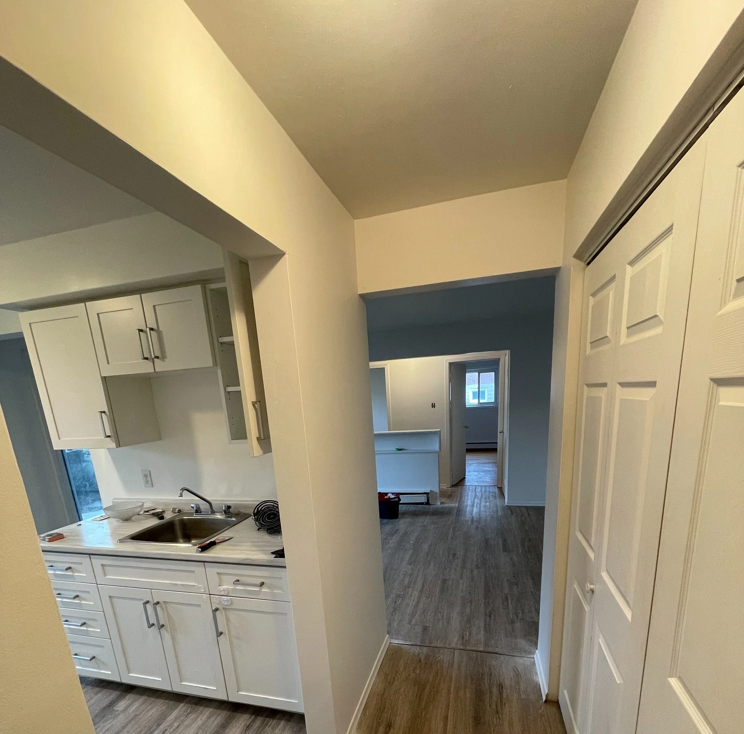 Interior view of a kitchen and living area under construction or renovation, with white cabinets, an open doorway, and a room with a window visible in the background.