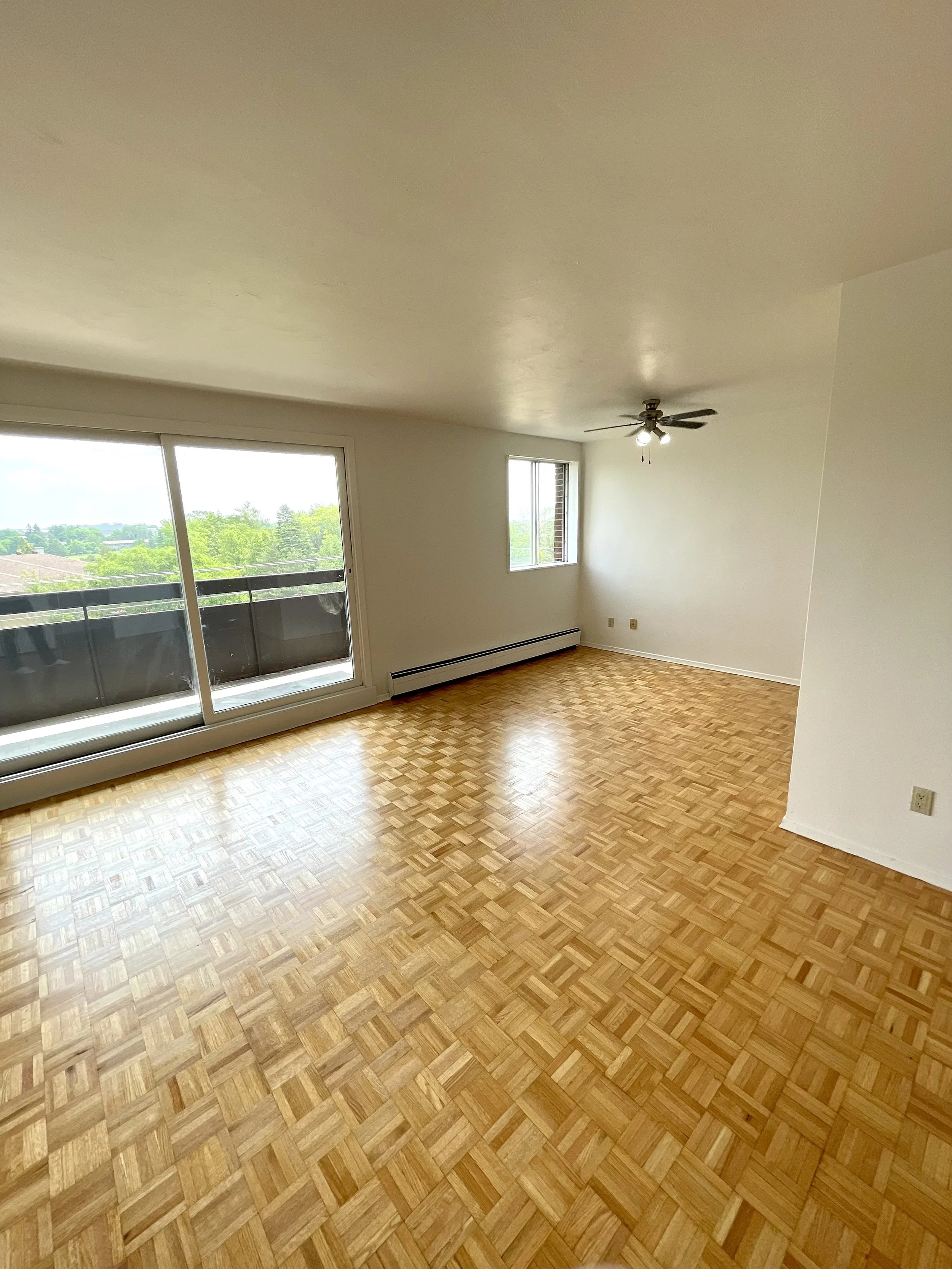 Empty living room with hardwood parquet flooring, sliding glass door leading to balcony, window, white walls, ceiling fan, and baseboard heater
