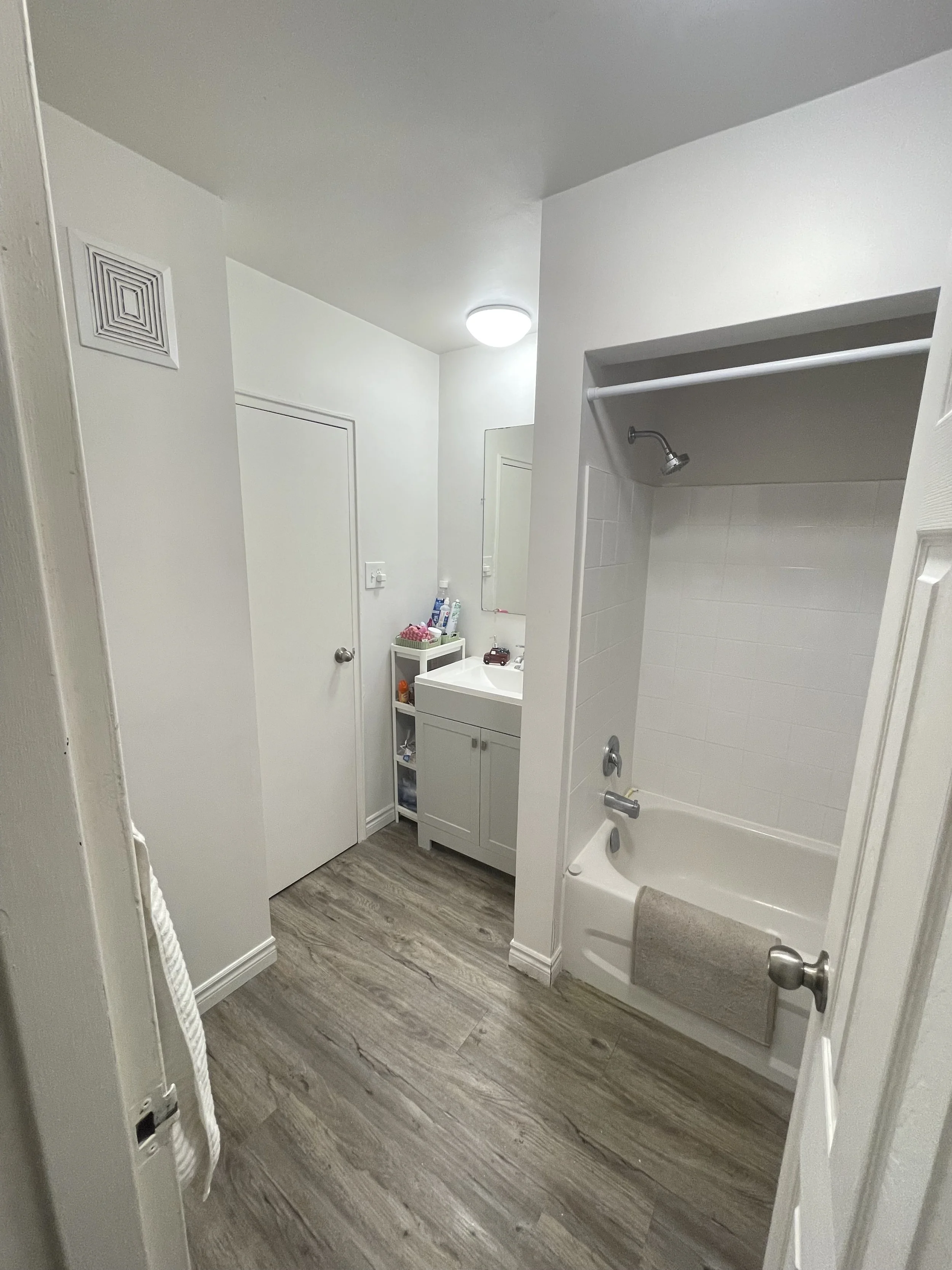 Bathroom with bathtub and shower, white walls, small vanity with sink, and wooden flooring.