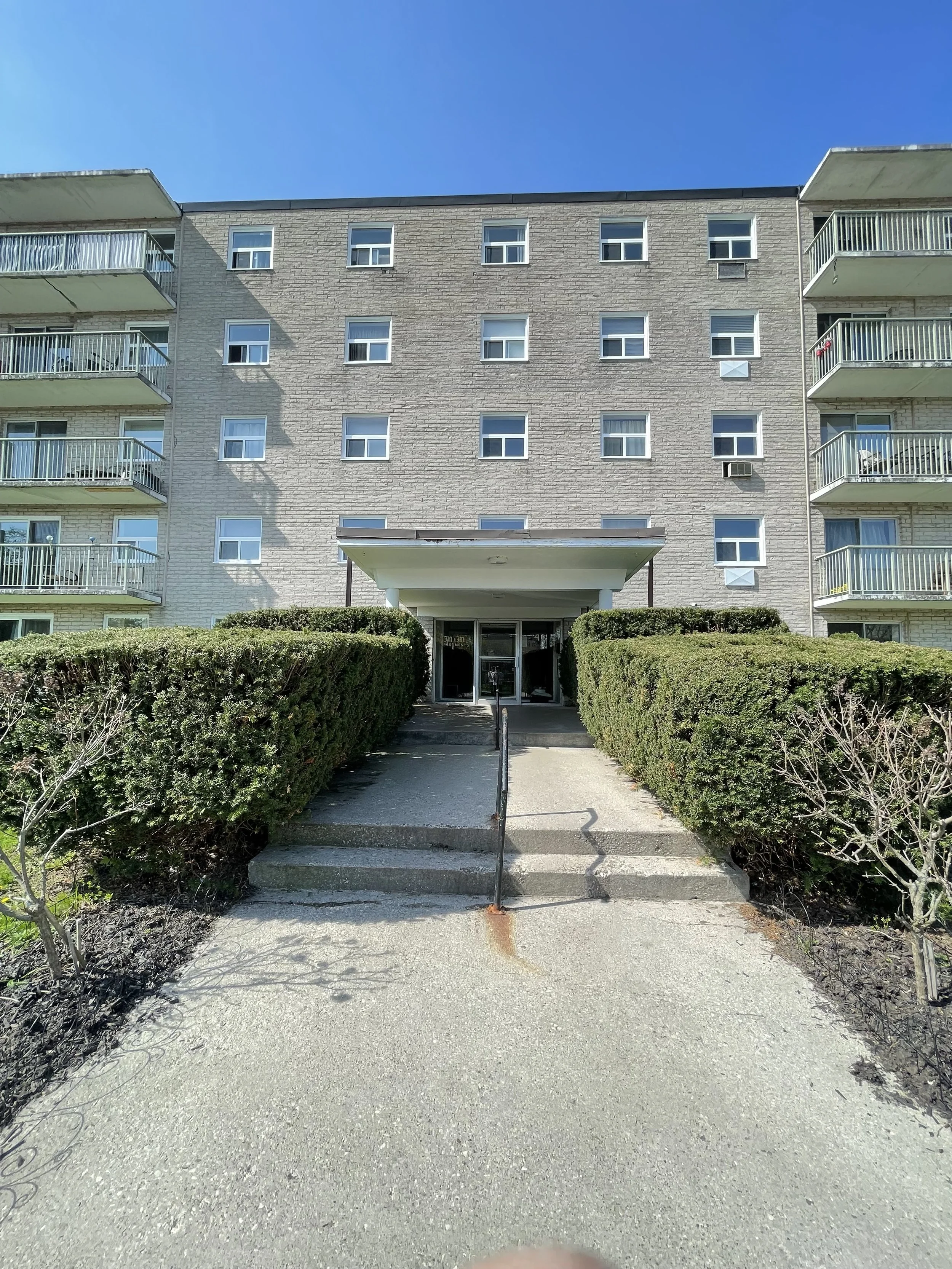 Entrance to a multi-story residential apartment building with balconies on both sides, a concrete walkway, green bushes, and a clear blue sky.