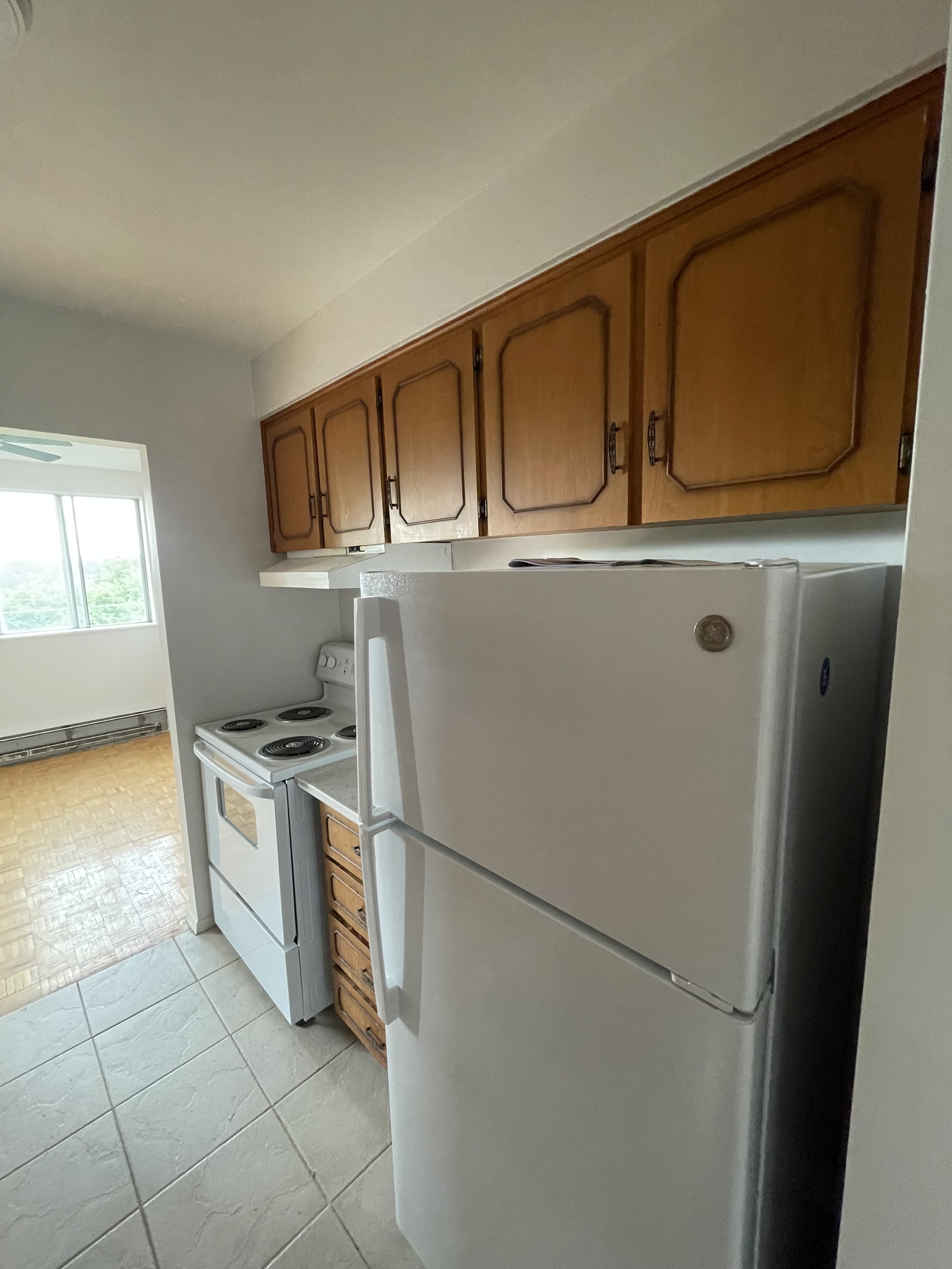 Kitchen with white refrigerator, stove, wooden cabinets, tile floor, and a view of a brightly lit room with large window.