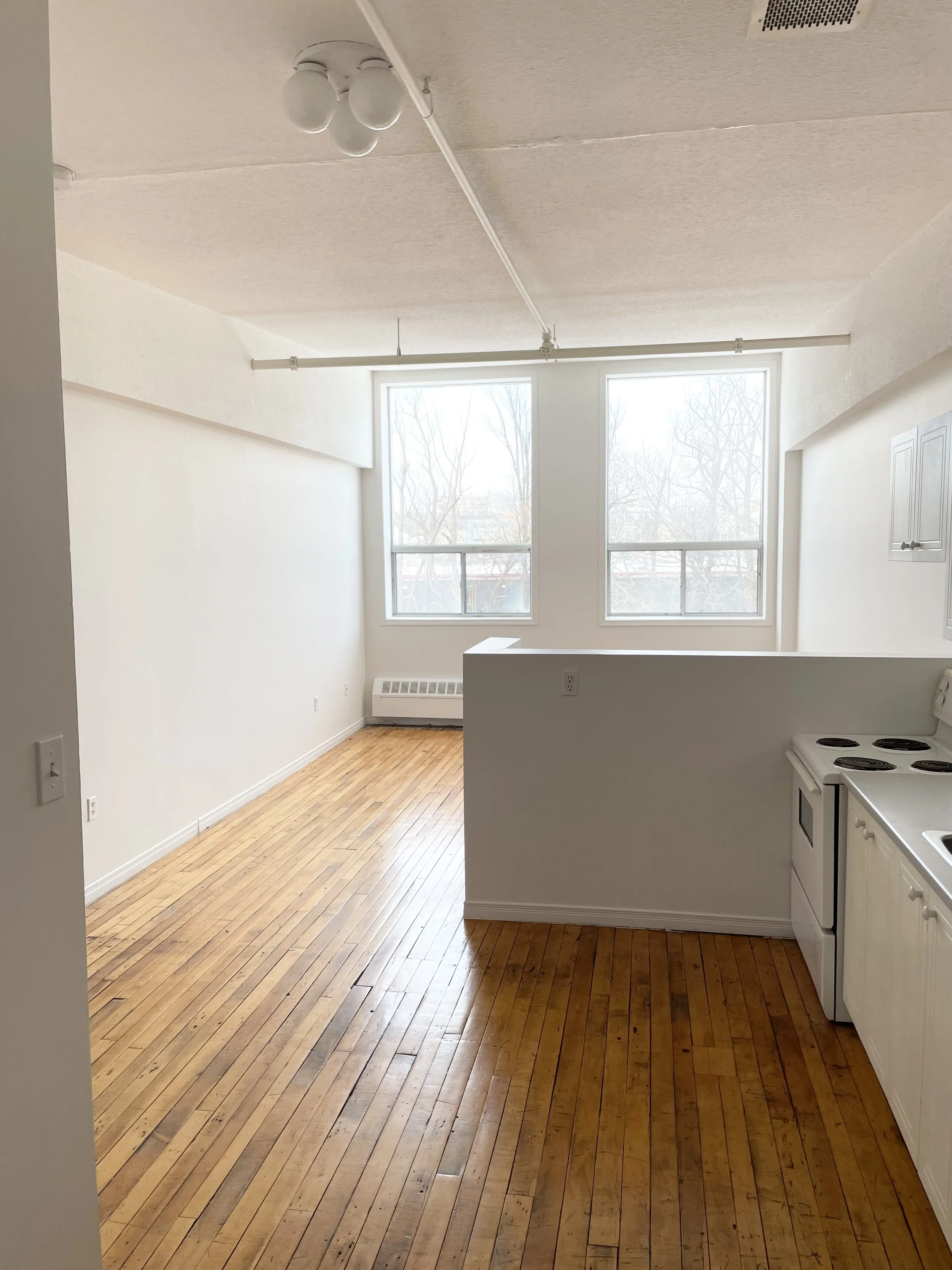 Empty apartment room with hardwood floors, white walls, large windows, and a small kitchen area with a stove and cabinets.