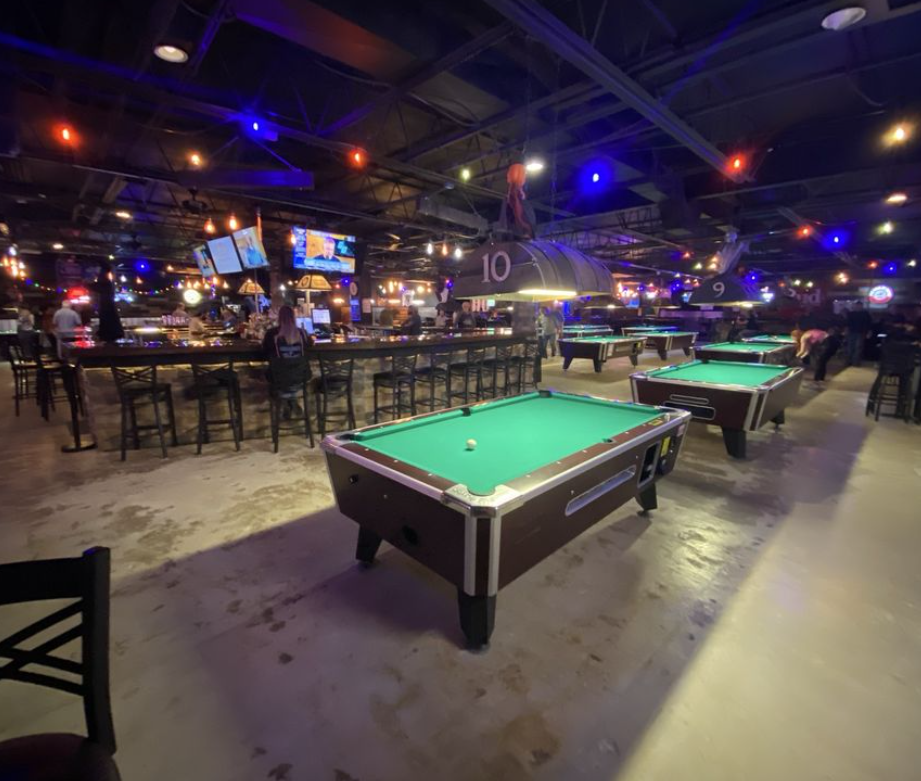 An indoor billiards and bar area with pool tables, bar seating, and multiple TV screens hanging from the ceiling. The area is decorated with colorful string lights and has a lively, energetic atmosphere at Miami Valley Sports Bar