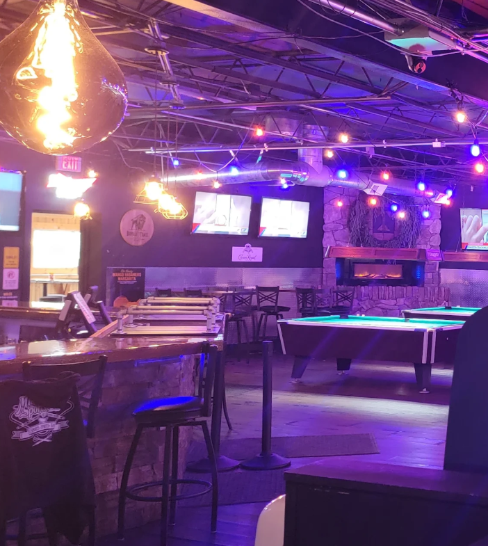 Interior of Miami Valley Sports Bar with pool tables, bar stools, and multiple televisions on the wall, illuminated by colorful string lights and vintage-style hanging lamps.
