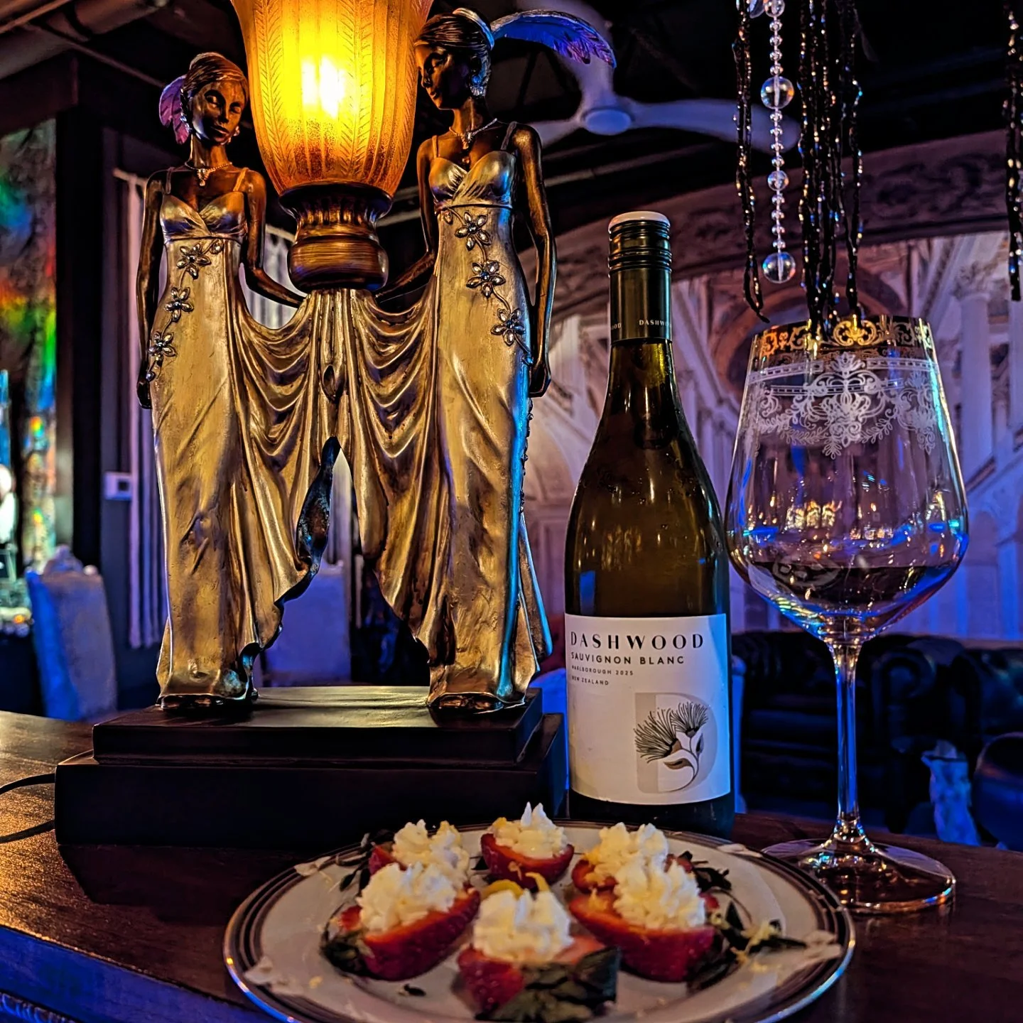 Decorative golden statue of two women holding a lamp, a bottle of Dashwood Sauvignon Blanc wine, a partly filled wine glass with decorative etching, and a plate of strawberries filled with cream on a table in a dimly lit, elegant setting.