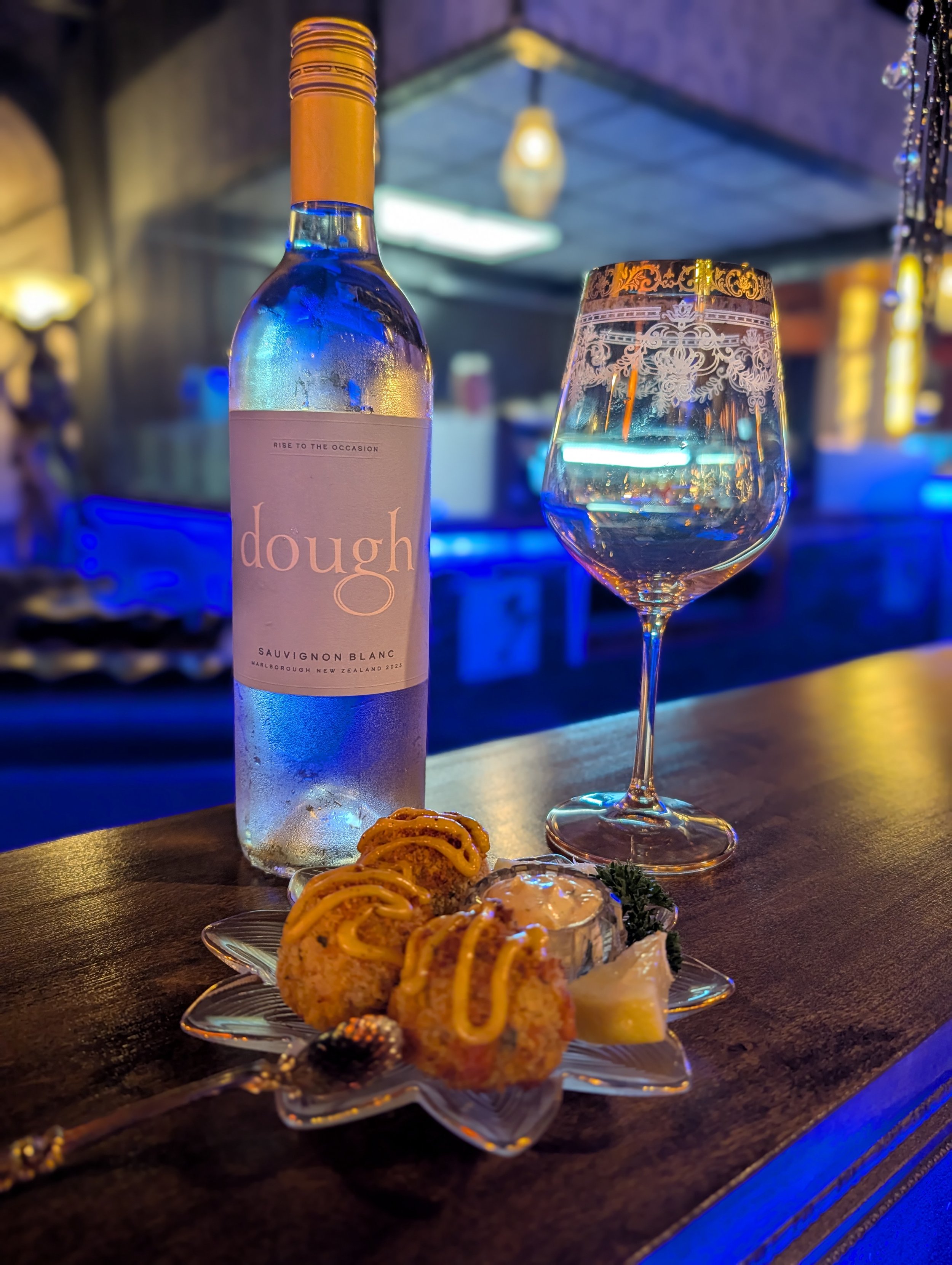 A bottle of wine and a glass of white wine on a bar counter, with a delectable small plate at Whisperz Speakeasy.