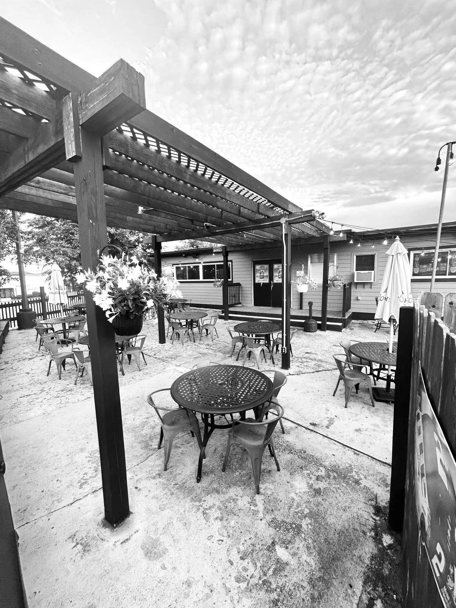 Black and white photo of an outdoor patio area with round metal tables and chairs, a wooden pergola structure overhead, string lights, a building with windows and a door, potted plants, and a street lamp in the background.
