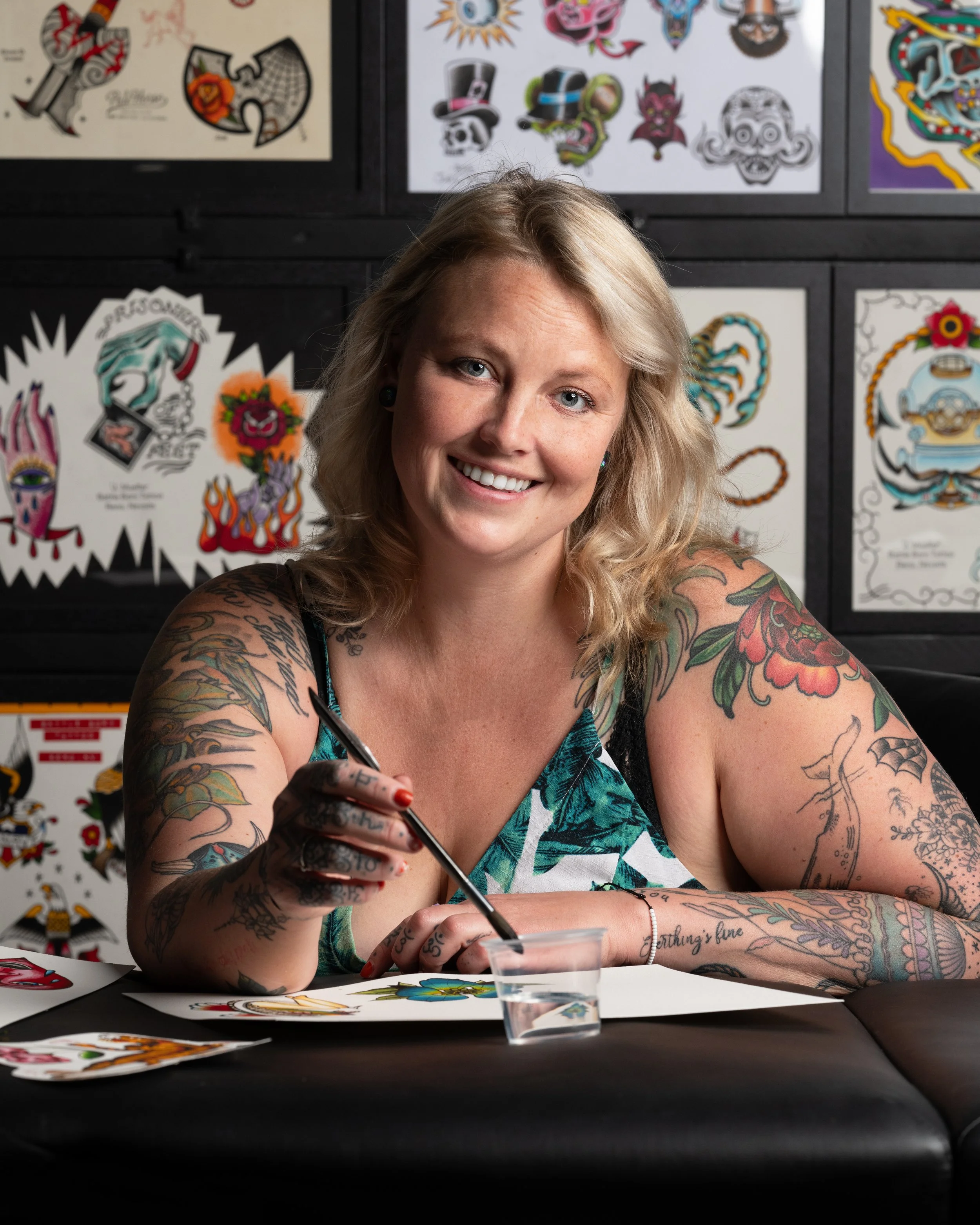 A woman with blonde hair and tattoos on her arms smiling at the camera while signing the artwork in front of her at an art event or convention. Behind her are display boards with various tattoo-style artwork and designs.
