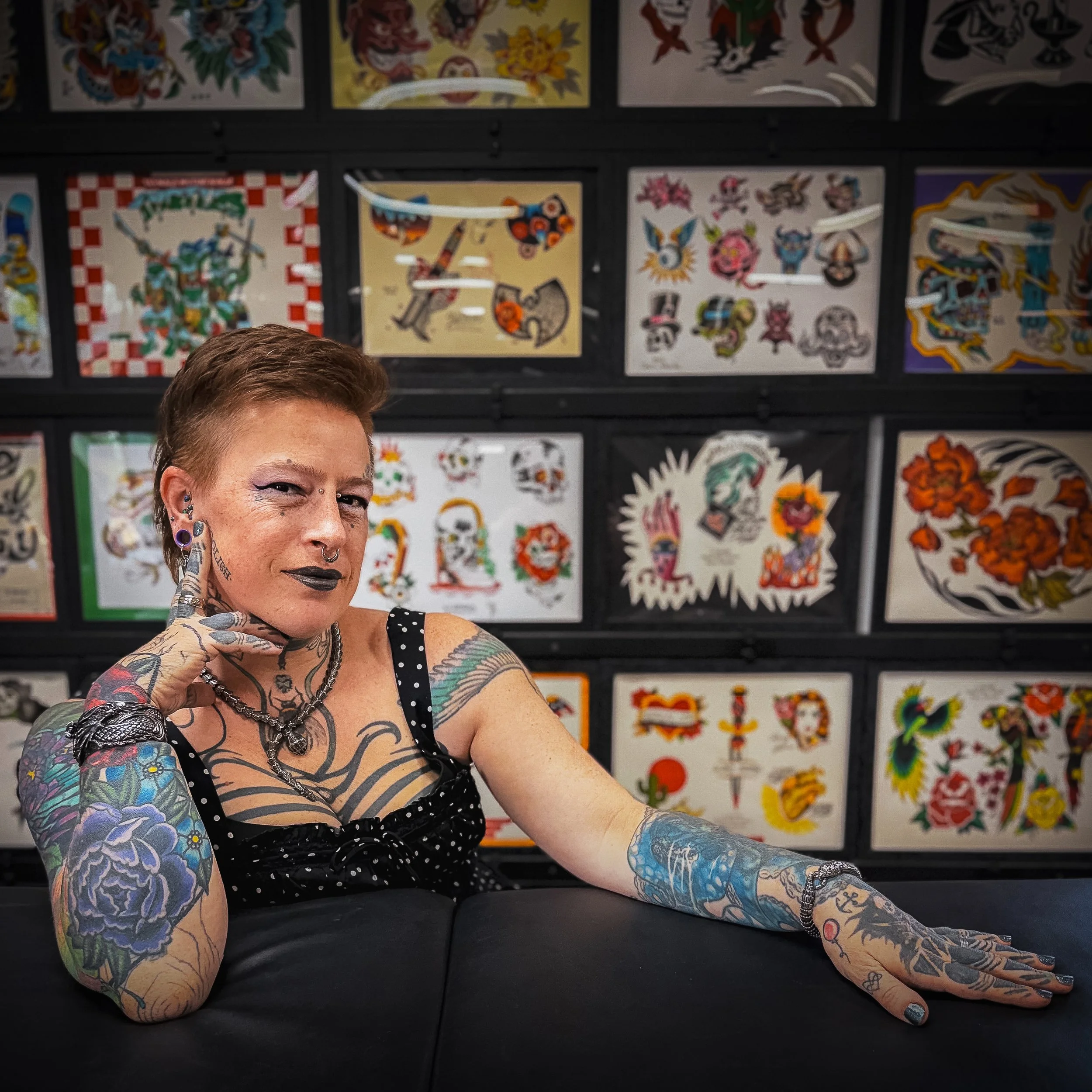 A person with tattoos and earrings smiling at the camera, with tattooed arms and neck, sitting in front of a wall decorated with colorful tattoo flash art designs.