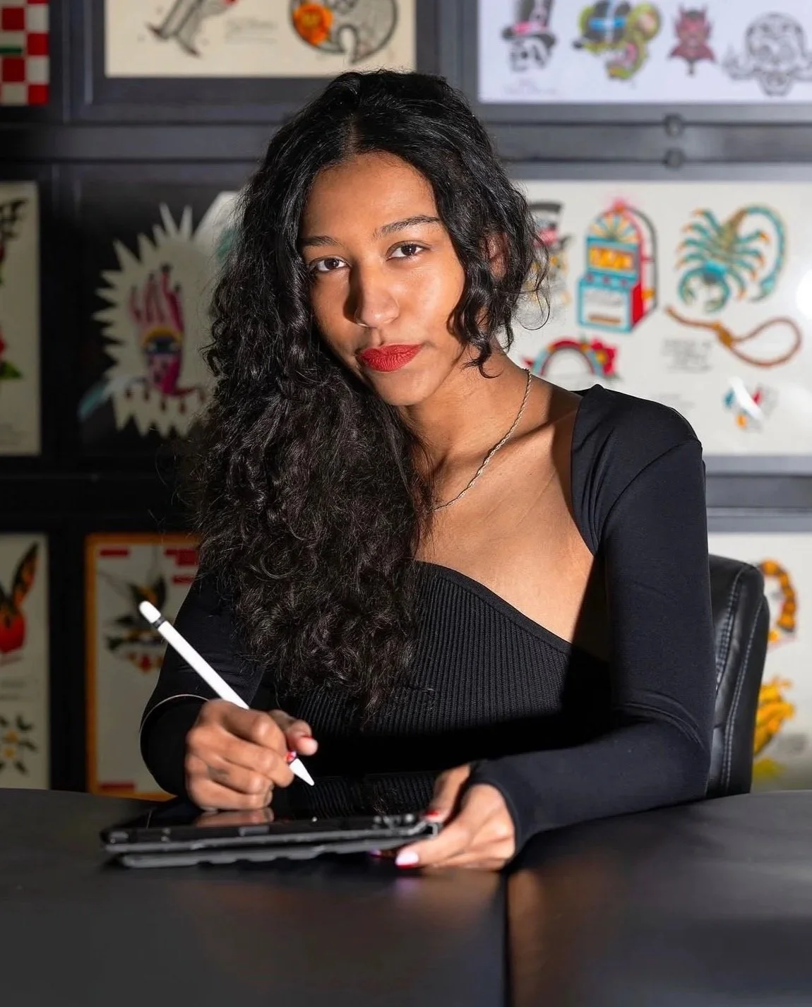 A woman with curly black hair and red lipstick is sitting at a table with a tablet and stylus. She is wearing a black top and a silver necklace, and is looking at the camera with a slight smile. The background features a display of colorful tattoo flash art on a wall.